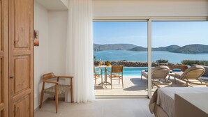 Una habitación con cama, un armario de madera, una silla, una mesa con una botella y fruta, y vistas al mar y a las montañas a través de un ventanal.