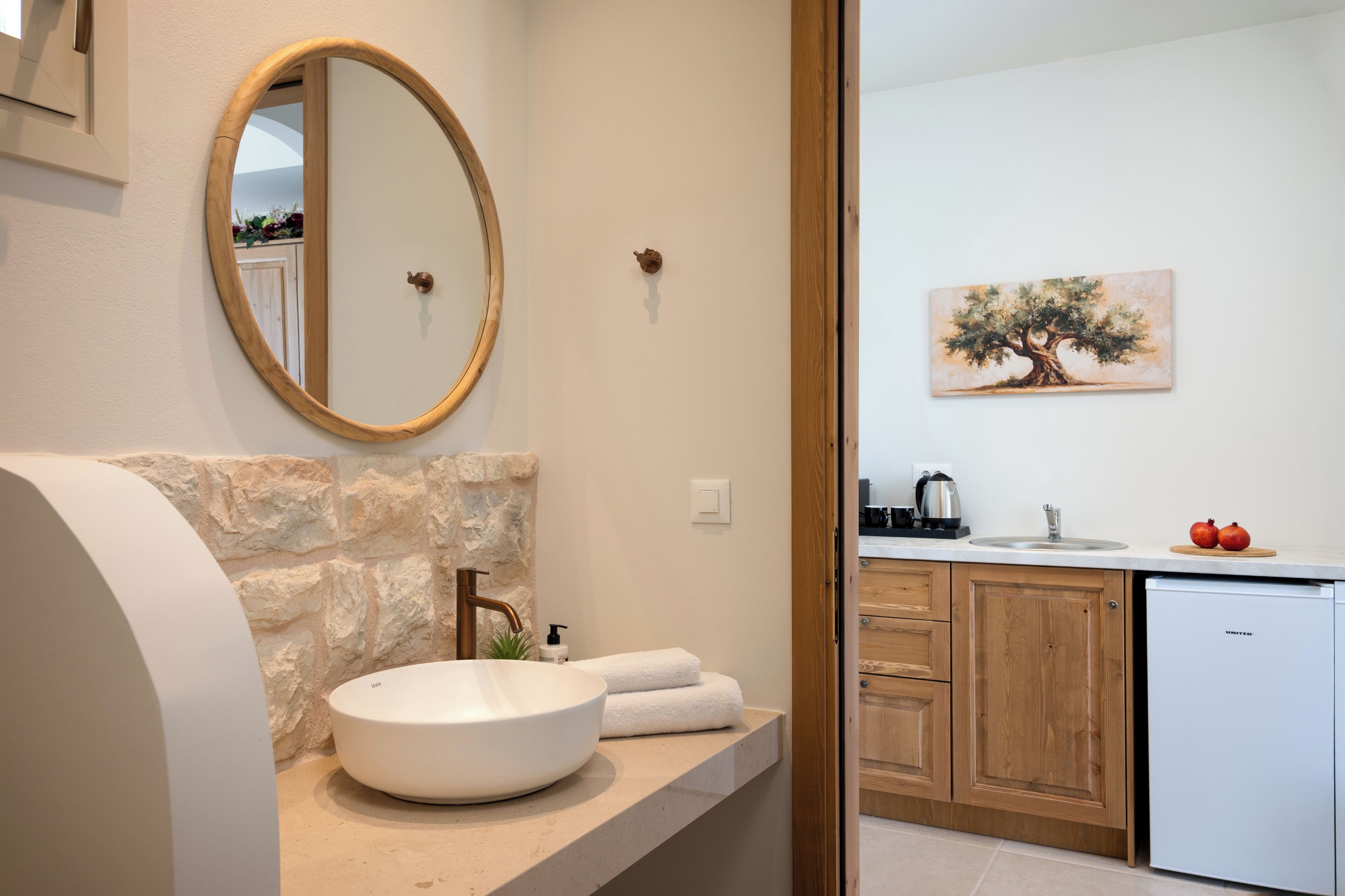 A bathroom with a round mirror, a white sink, a stone wall, a towel, and a painting of a tree.