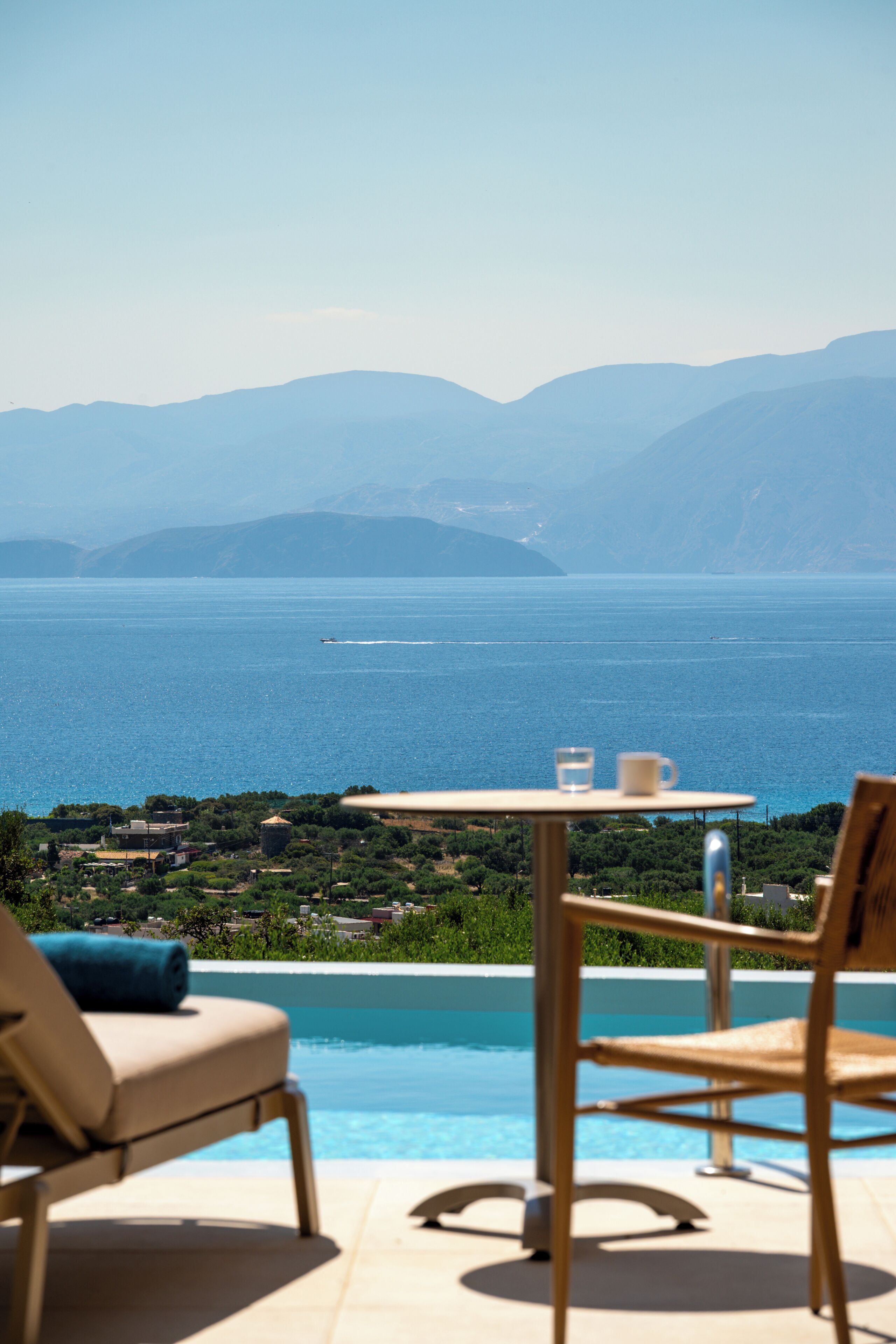 A poolside view with a table and chairs, overlooking a large body of water and distant mountains.