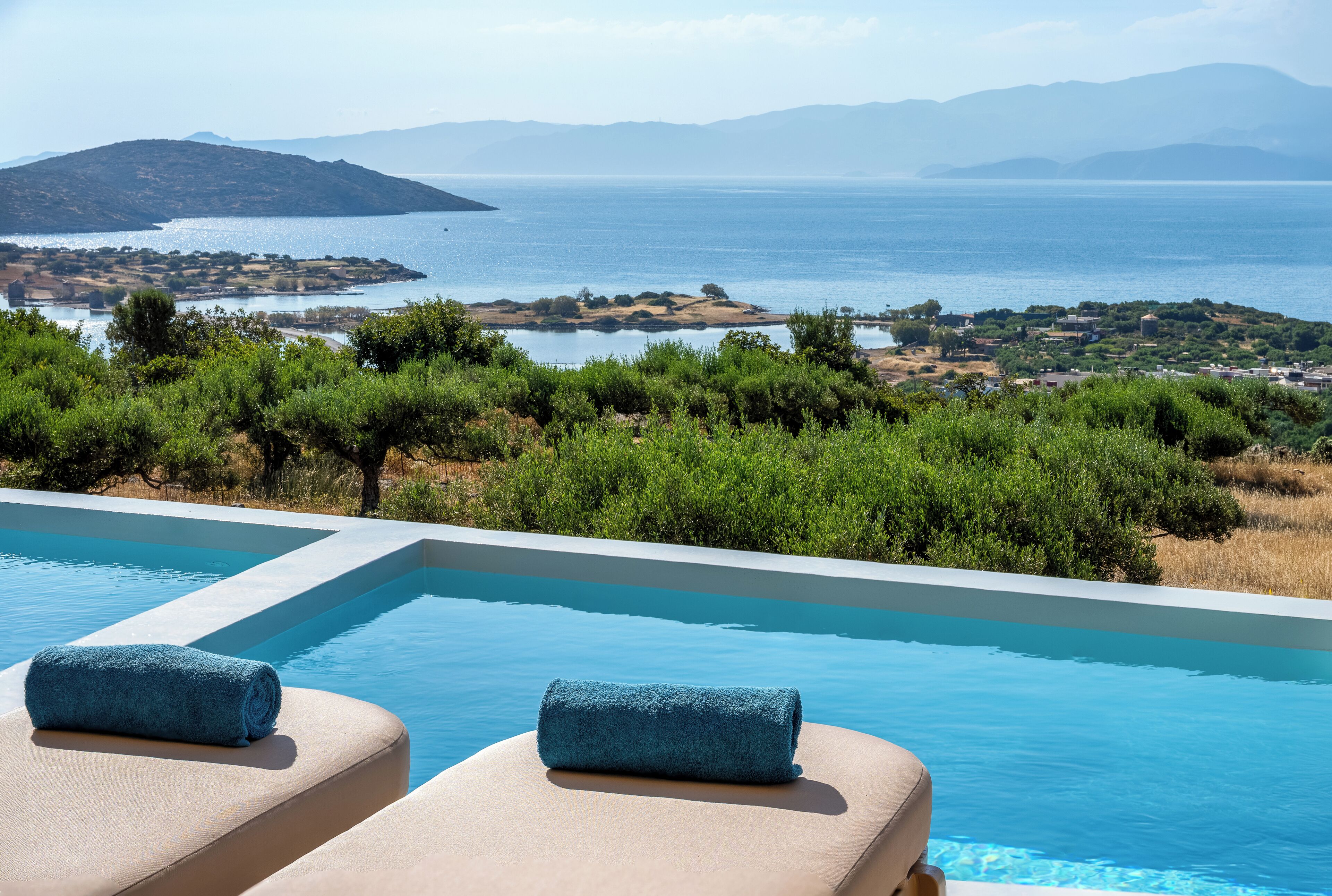 A pool with two lounge chairs, a towel, and a view of the sea and hills.