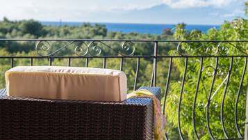 A wicker chair with a beige cushion and a colorful towel on a balcony overlooking a lush green landscape and the ocean.