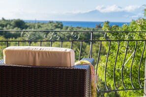 Una silla de mimbre con un cojín color crema y una toalla de colores en un balcón con vistas a un paisaje verde exuberante y al océano.
