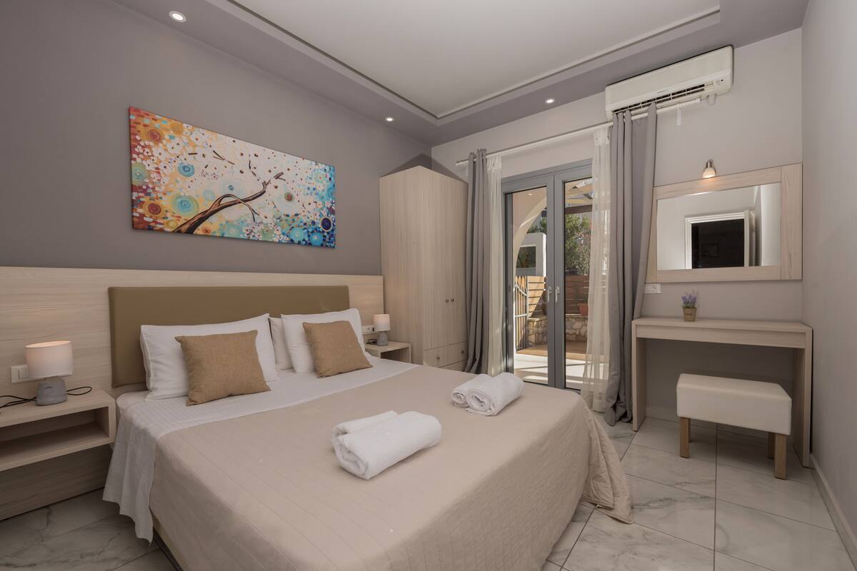 A modern bedroom with a large bed, a nightstand, a desk, a mirror, and a sliding glass door leading to an outdoor area.