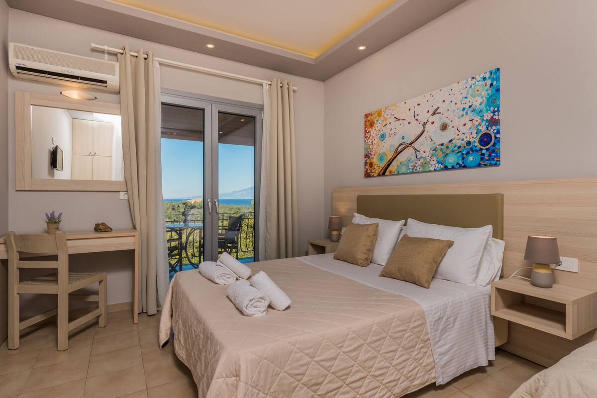 A hotel room with a bed, a desk, a chair, a nightstand, a lamp, a painting, and a view of the outdoors.