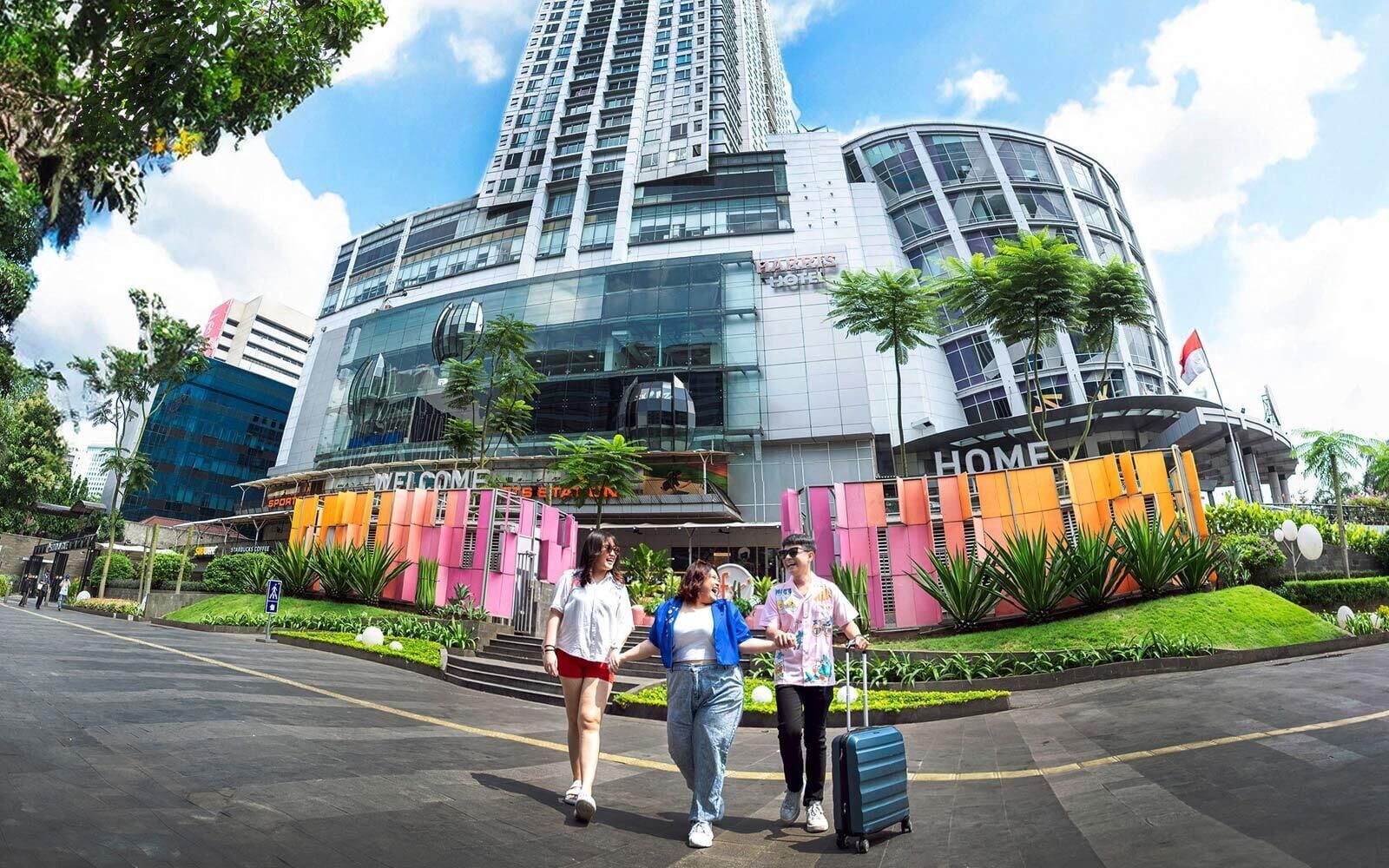 A modern hotel with a curved glass facade, a prominent entrance with a 'Welcome' sign, and a well-maintained garden area.