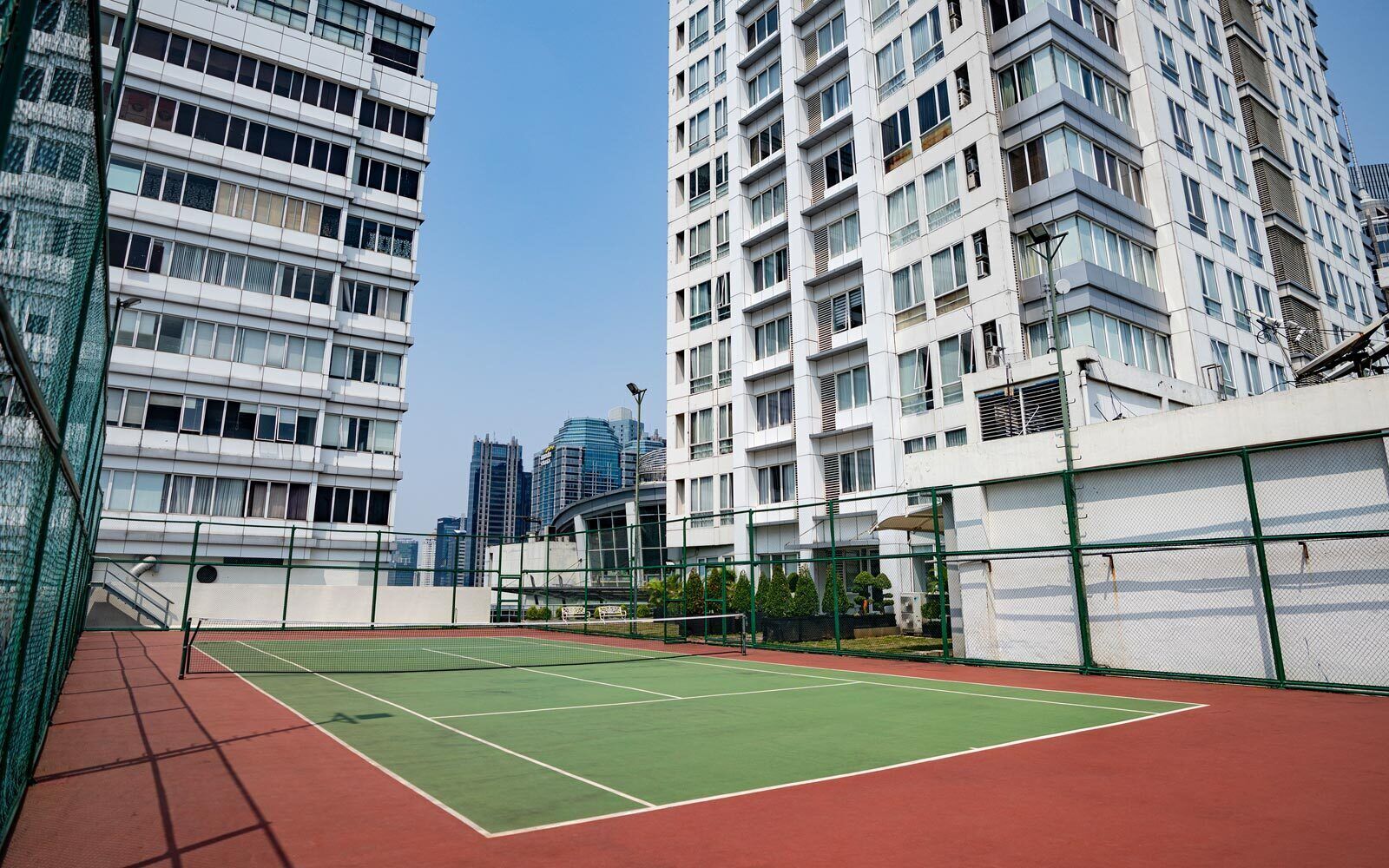 A tennis court with a net, surrounded by a green fence, and adjacent to a high-rise building with multiple windows.