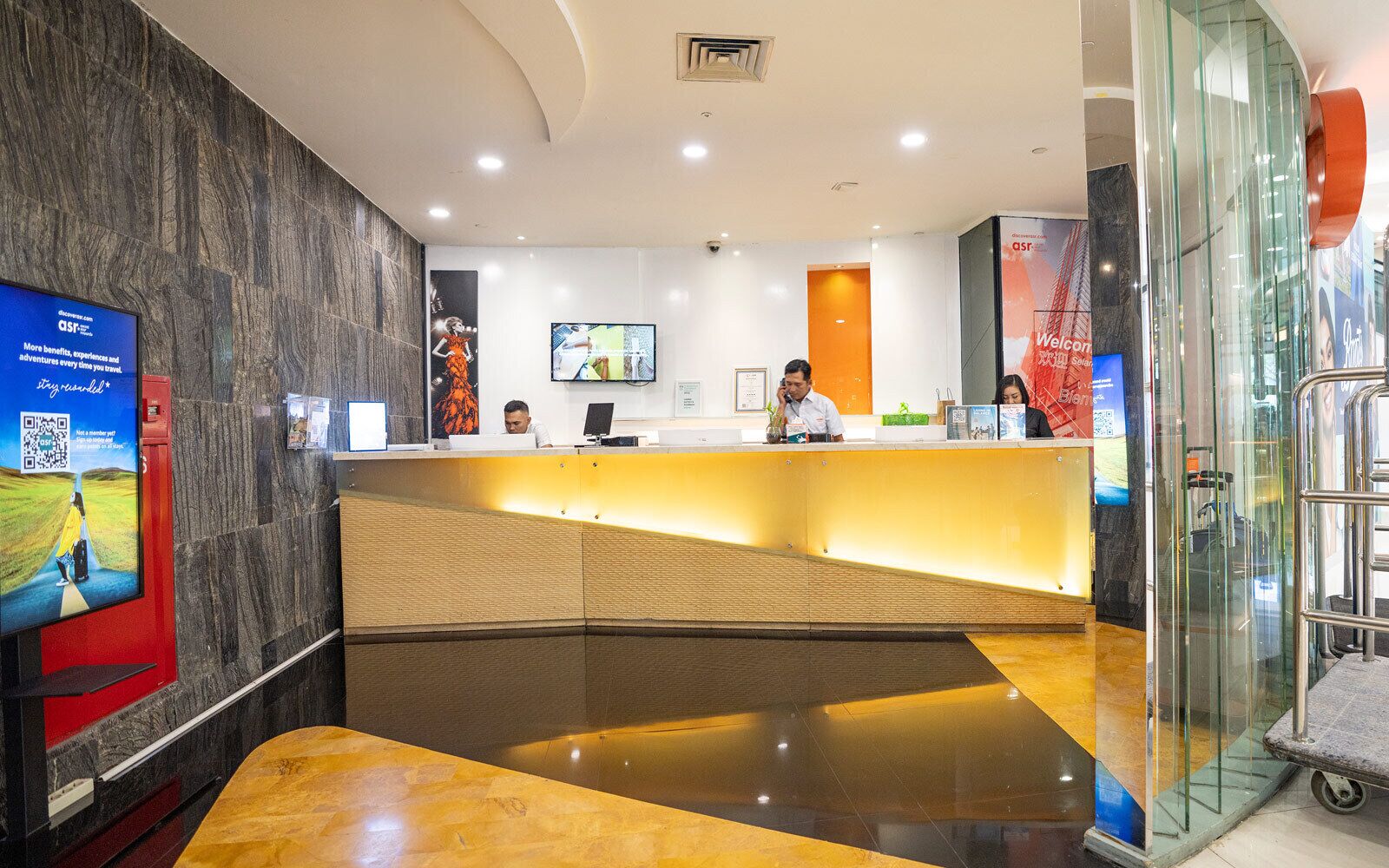 En hotellobby med modern design, en receptionsdisk, en digital skärm och en glashiss.