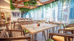 A restaurant with wooden tables and chairs, green hanging plants, and a city view through large windows. - HARRIS Suites FX Sudirman Jakarta (Jakarta)