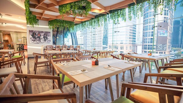 A restaurant with wooden tables and chairs, green hanging plants, and a city view through large windows. - HARRIS Suites FX Sudirman Jakarta (Jakarta)