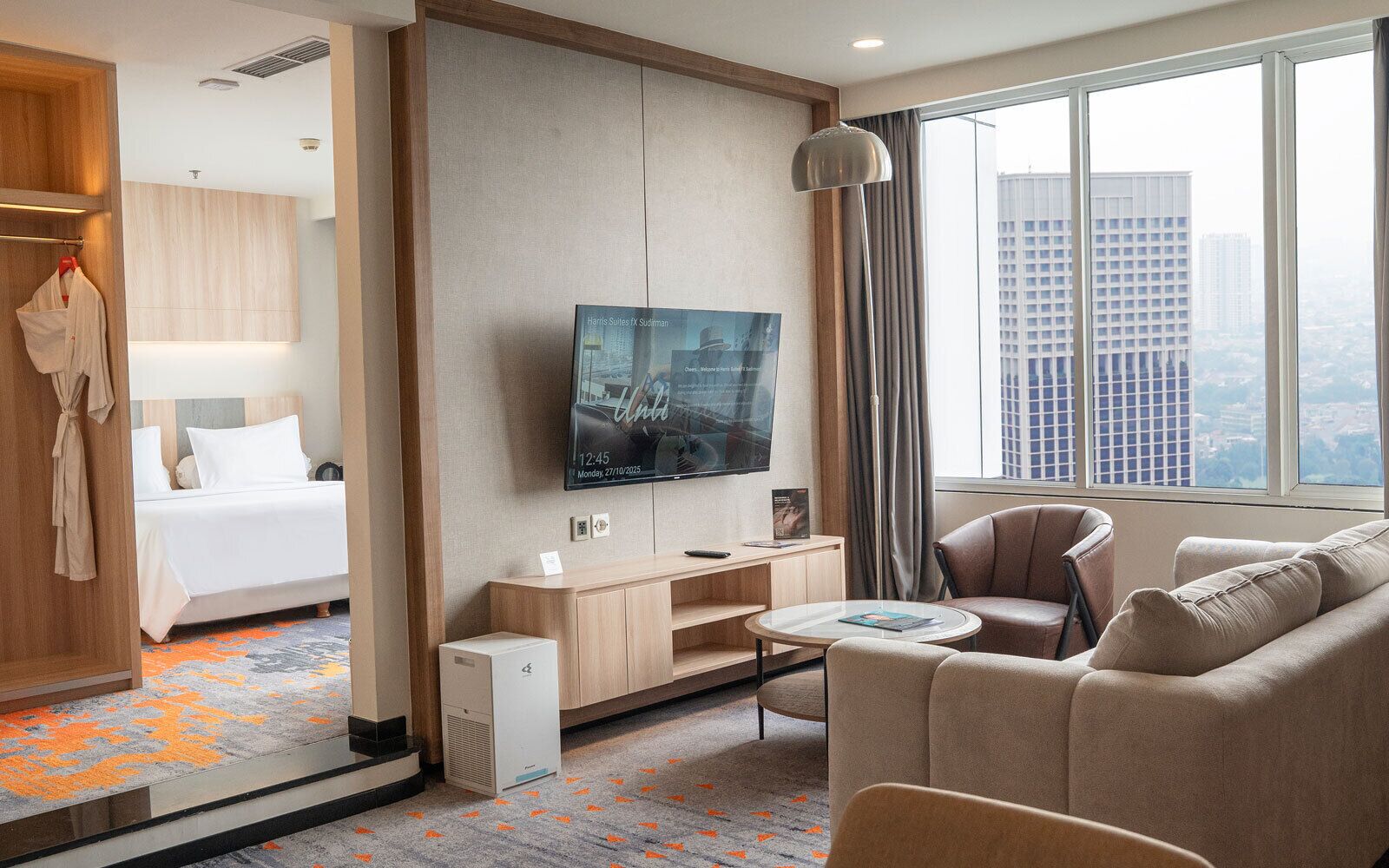A hotel room with a bed, a TV, a seating area with a sofa and armchairs, and a view of the city through large windows.