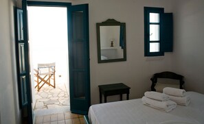 Quarto com cama, mesa de cabeceira, espelho e porta aberta para um pátio com uma cadeira.