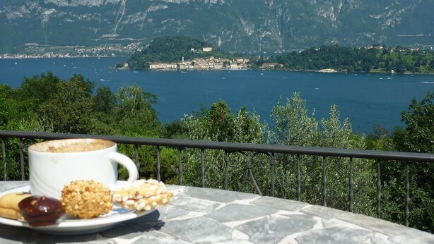 Una vista panorámica de un lago con montañas al fondo, una mesa con una taza de café y pasteles, y una barandilla.