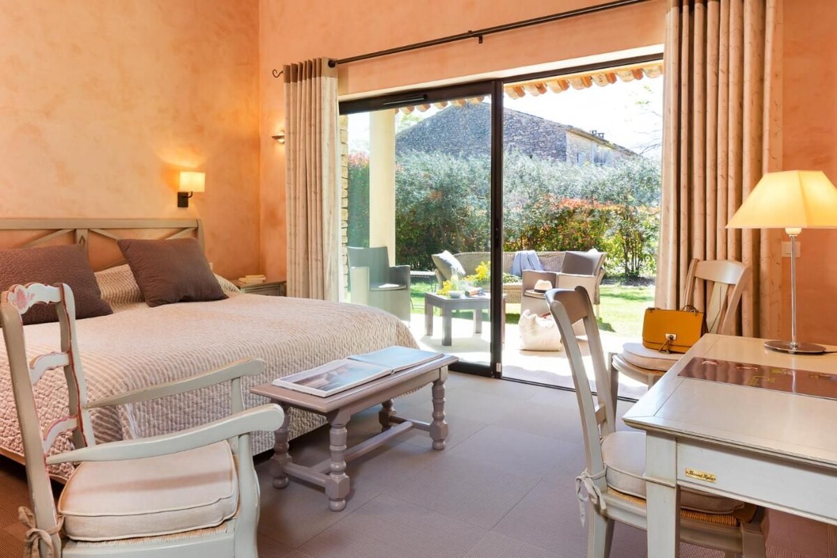 A bedroom with a bed, a desk, a chair, and a table. There is a window with curtains and a view of the outside.