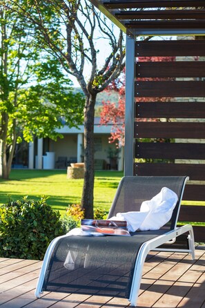 A sun lounger with a magazine, positioned on a wooden deck with a view of a garden and a building in the background. - Hotel Carcarille - Restaurant le C (Gordes)