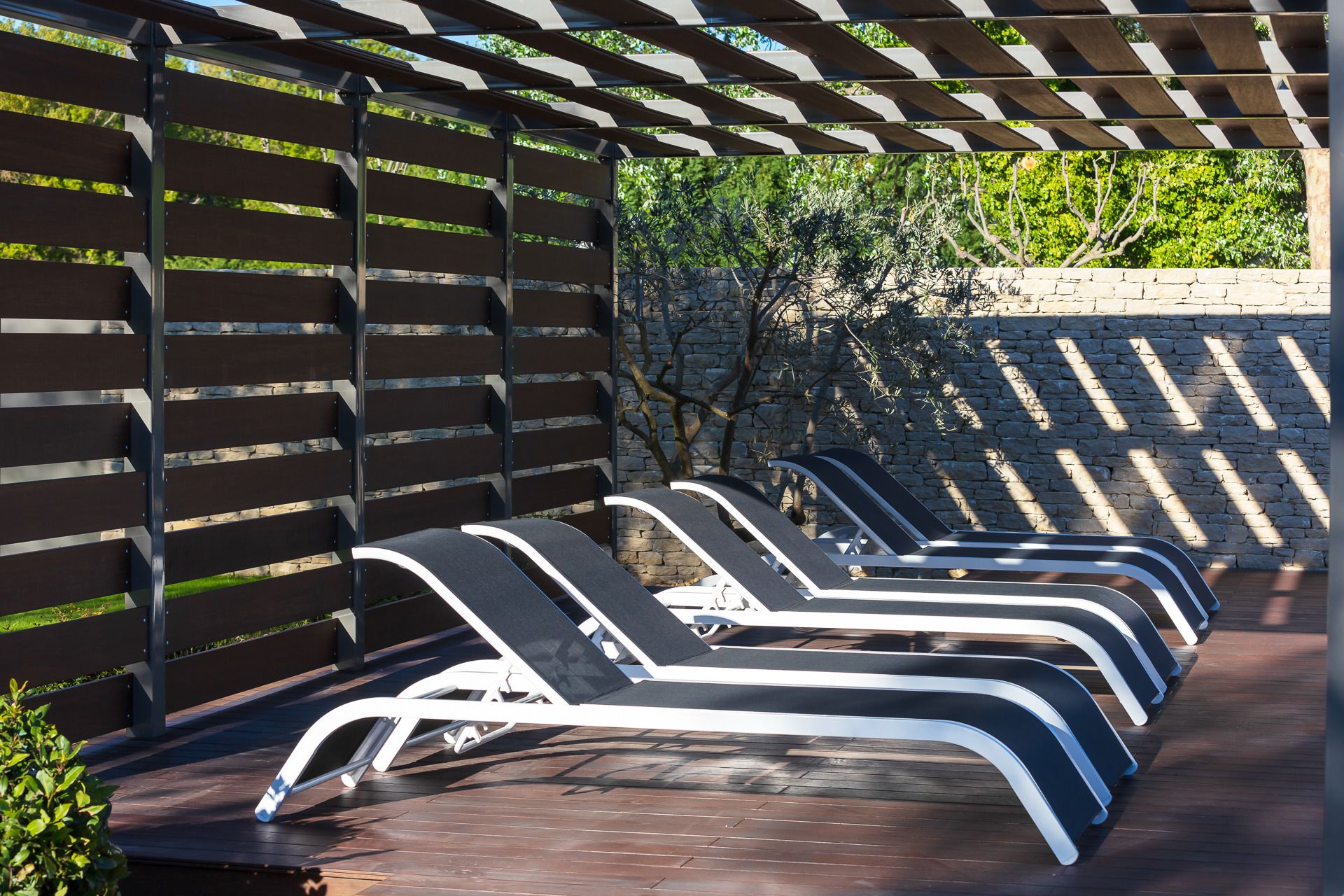 A row of sun loungers under a pergola with a stone wall and greenery in the background.