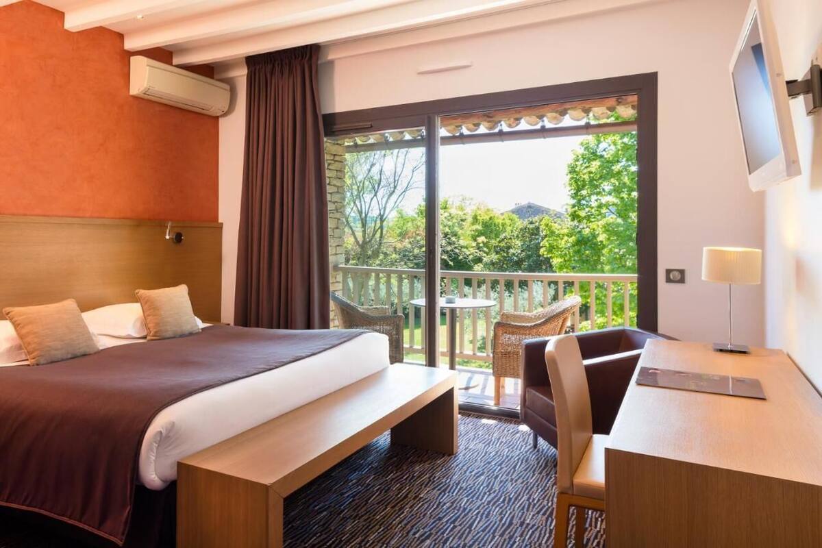 A hotel room with a large bed, a desk with a chair, a small balcony with wicker furniture, and a TV mounted on the wall.