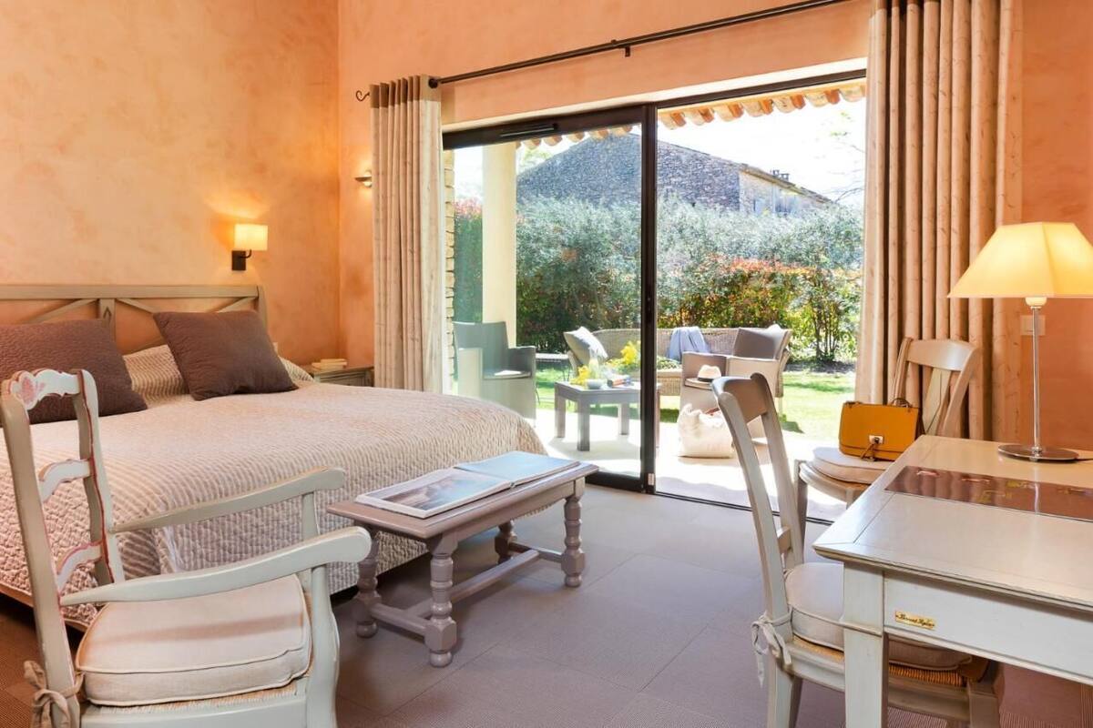 A bedroom with a bed, a desk, a chair, and a table. There is a window with curtains and a view of the outside.