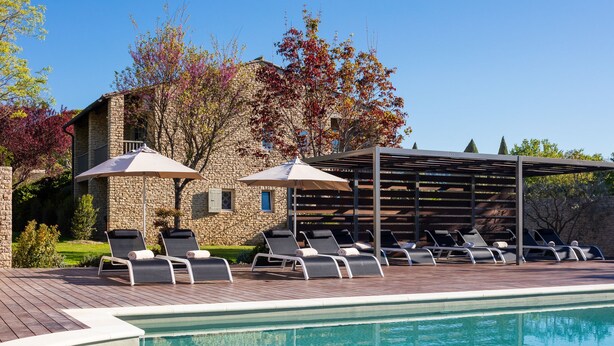 A pool area with lounge chairs and umbrellas, a stone building, and a pergola. - Hotel Carcarille - Restaurant le C (Gordes)