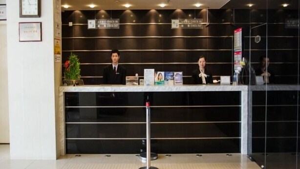 Un mostrador de recepción con dos empleados, un reloj y un cartel que indica las áreas de registro de entrada y salida.