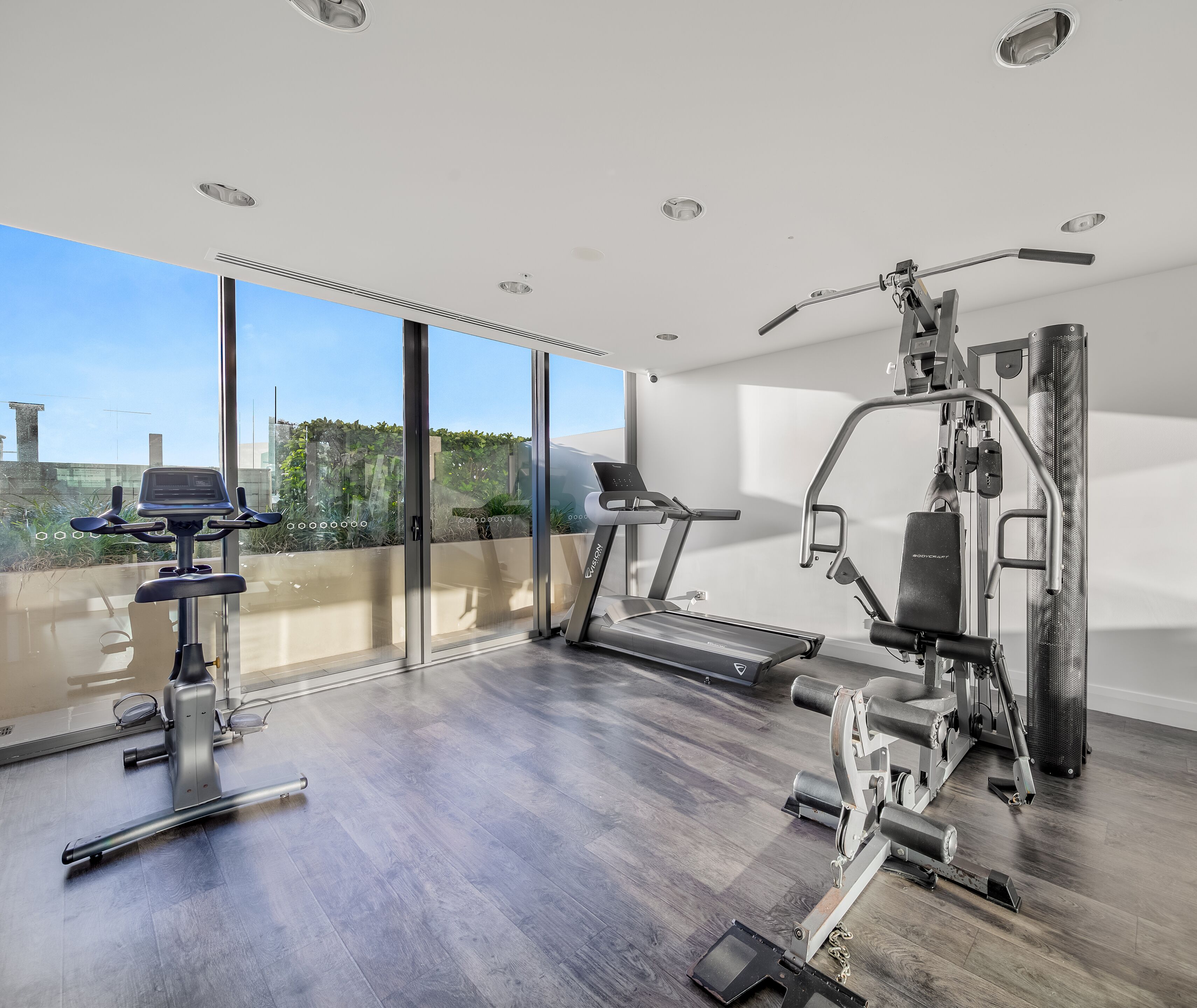 A modern home gym with a treadmill, elliptical, and weight machines, large windows, and a view of greenery.