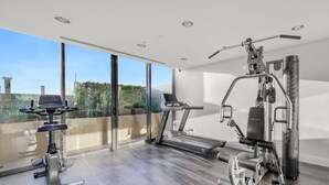A modern home gym with a treadmill, elliptical, and weight machines, large windows, and a view of greenery. - Oaks Redcliffe Mon Komo Suites (Redcliffe)