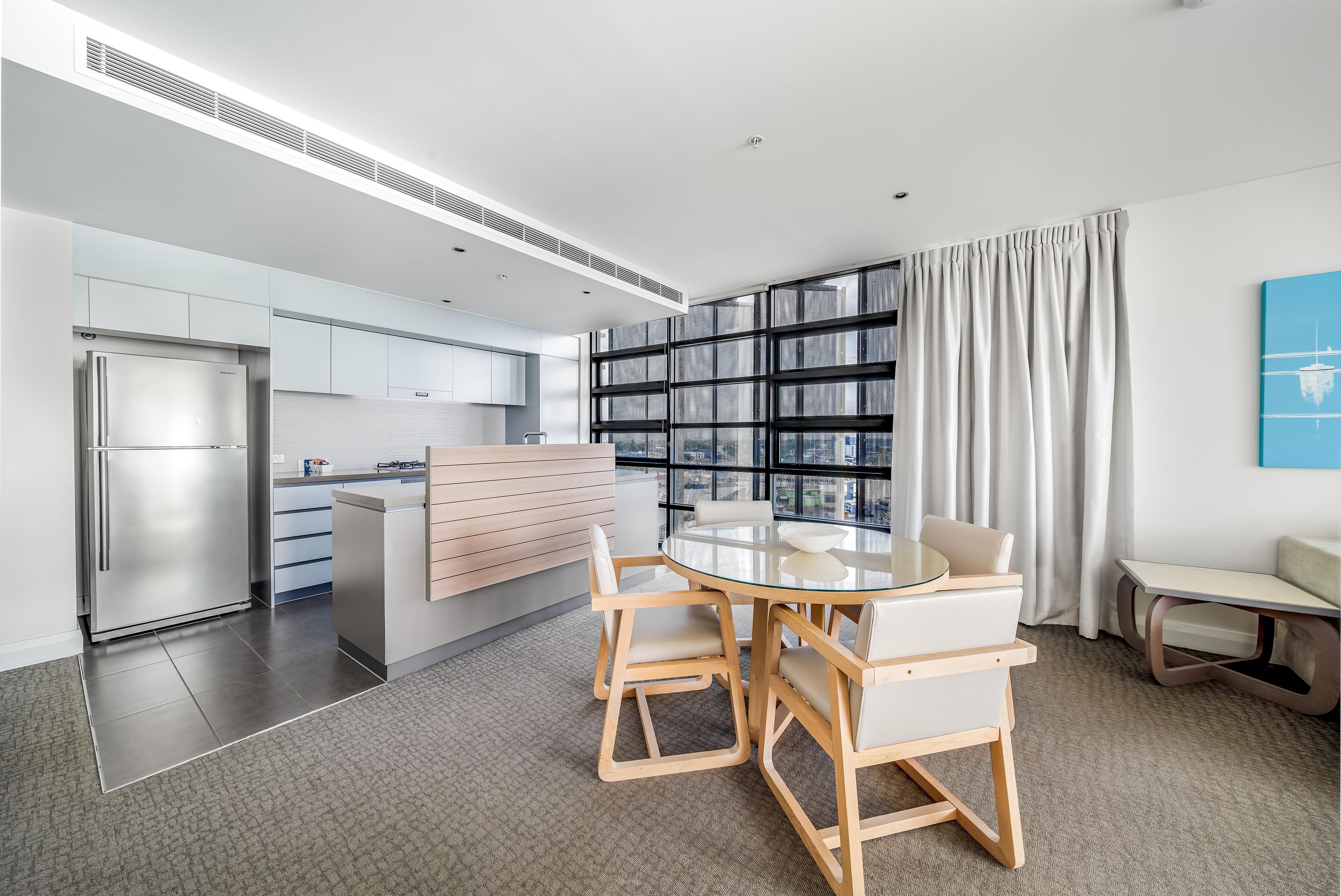 A modern kitchen with a built-in refrigerator, a dining area with a glass table and wooden chairs, and a view of the city through large windows.