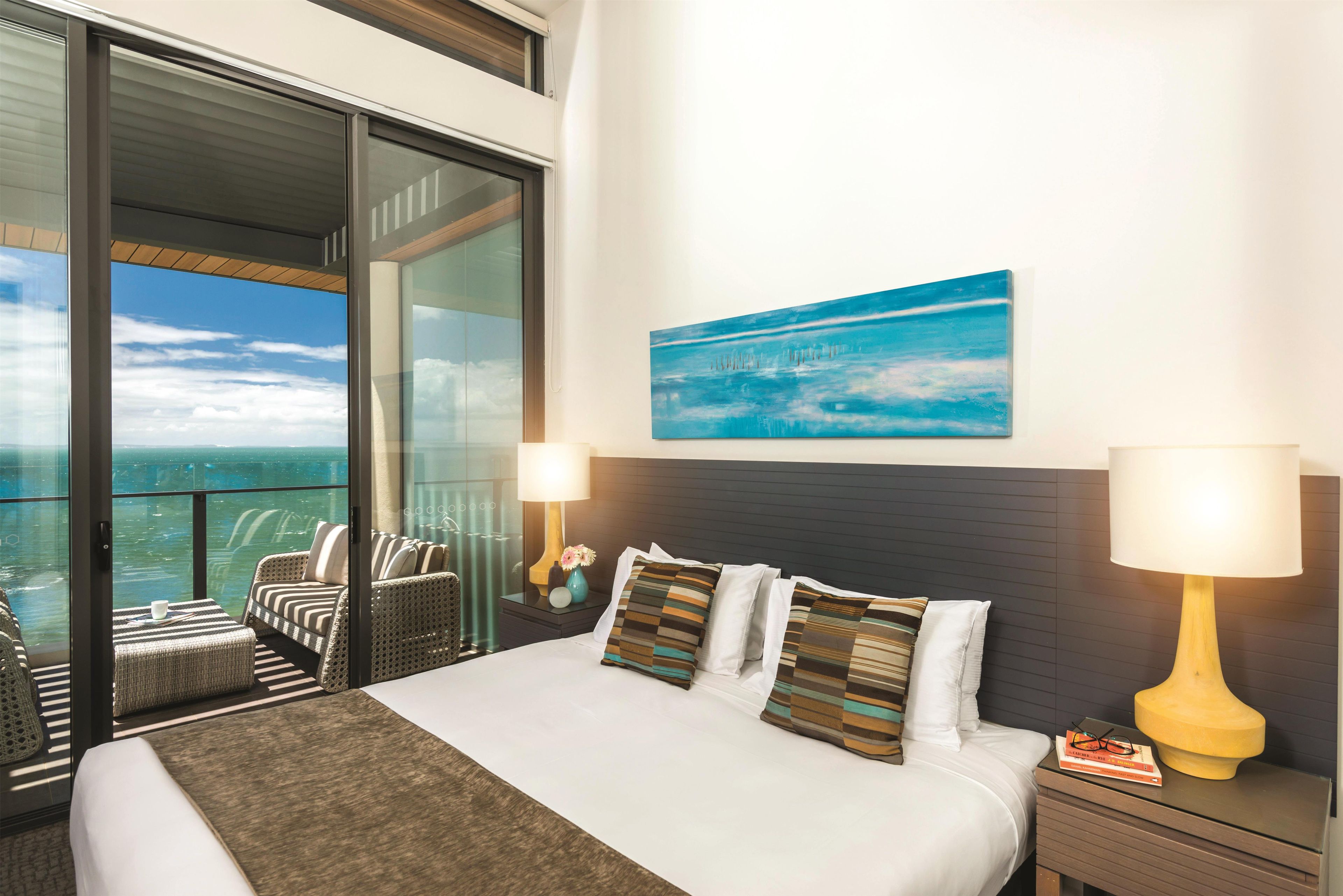 A hotel room with a bed, a nightstand, a lamp, a painting, and a view of the ocean.