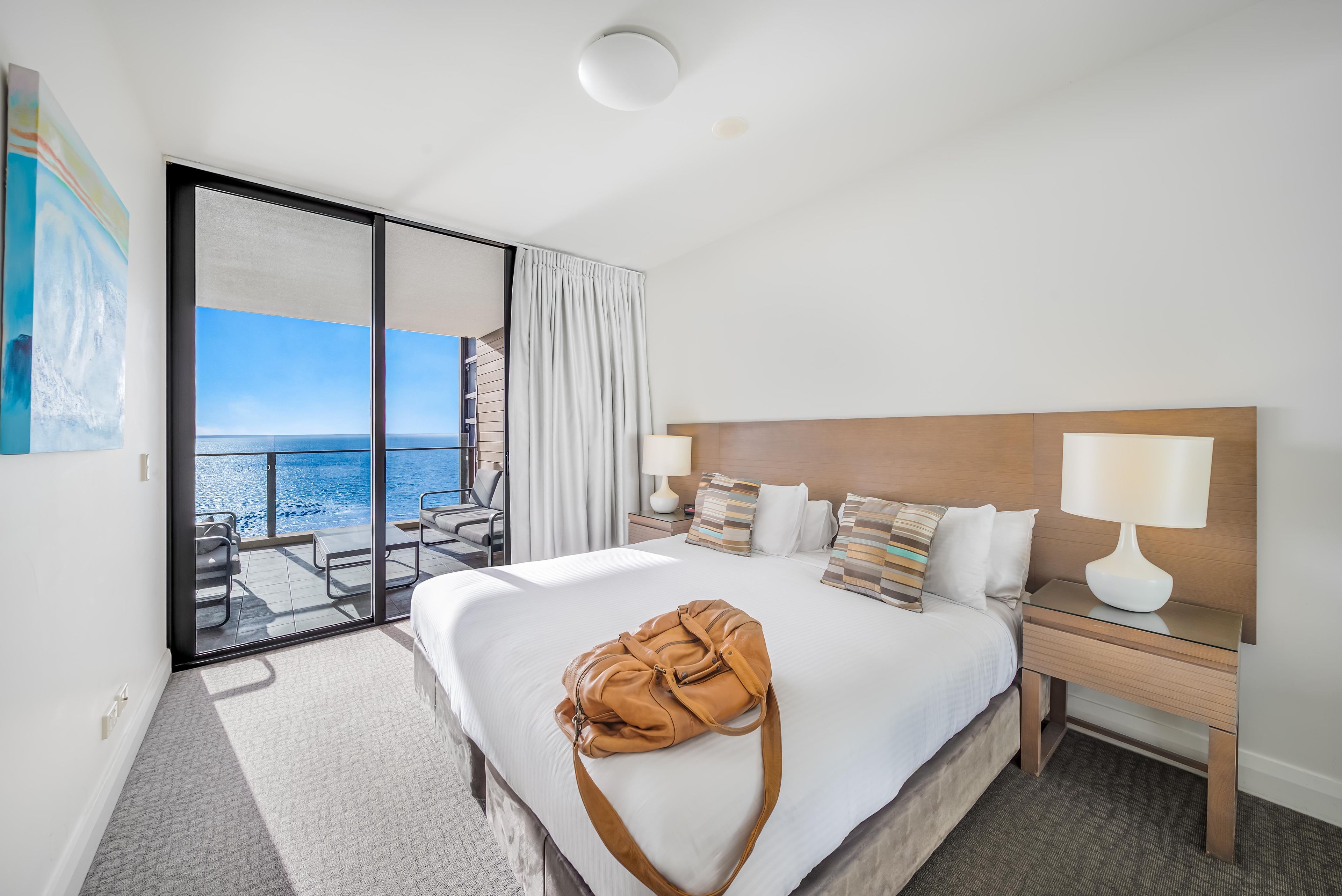 Une chambre d’hôtel avec un grand lit, une table de chevet, une lampe, une vue sur la mer et une porte coulissante donnant sur un balcon.