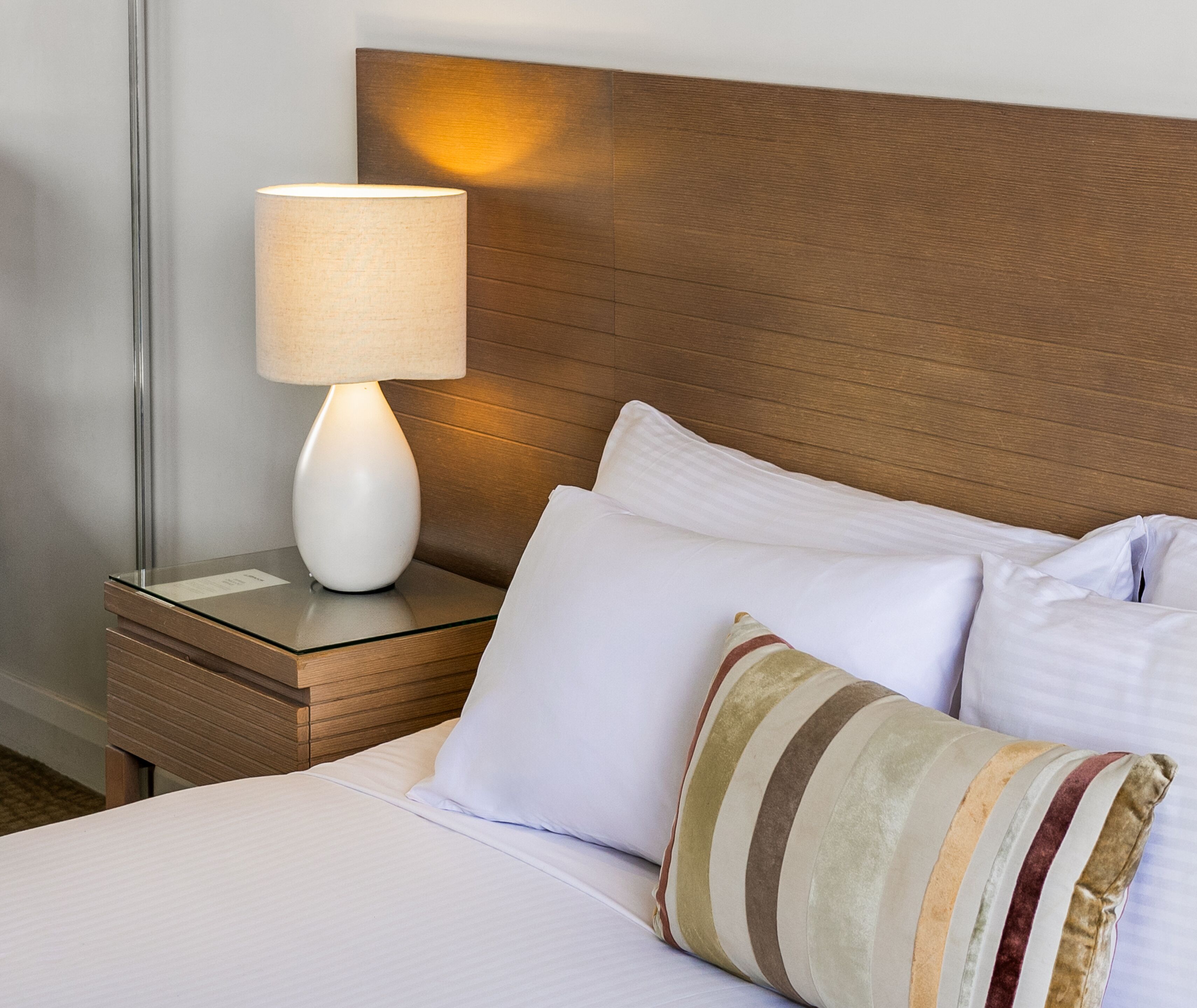 Un lit bien fait, avec du linge de lit blanc, un coussin à rayures et une lampe de chevet posée sur une table de nuit en bois.