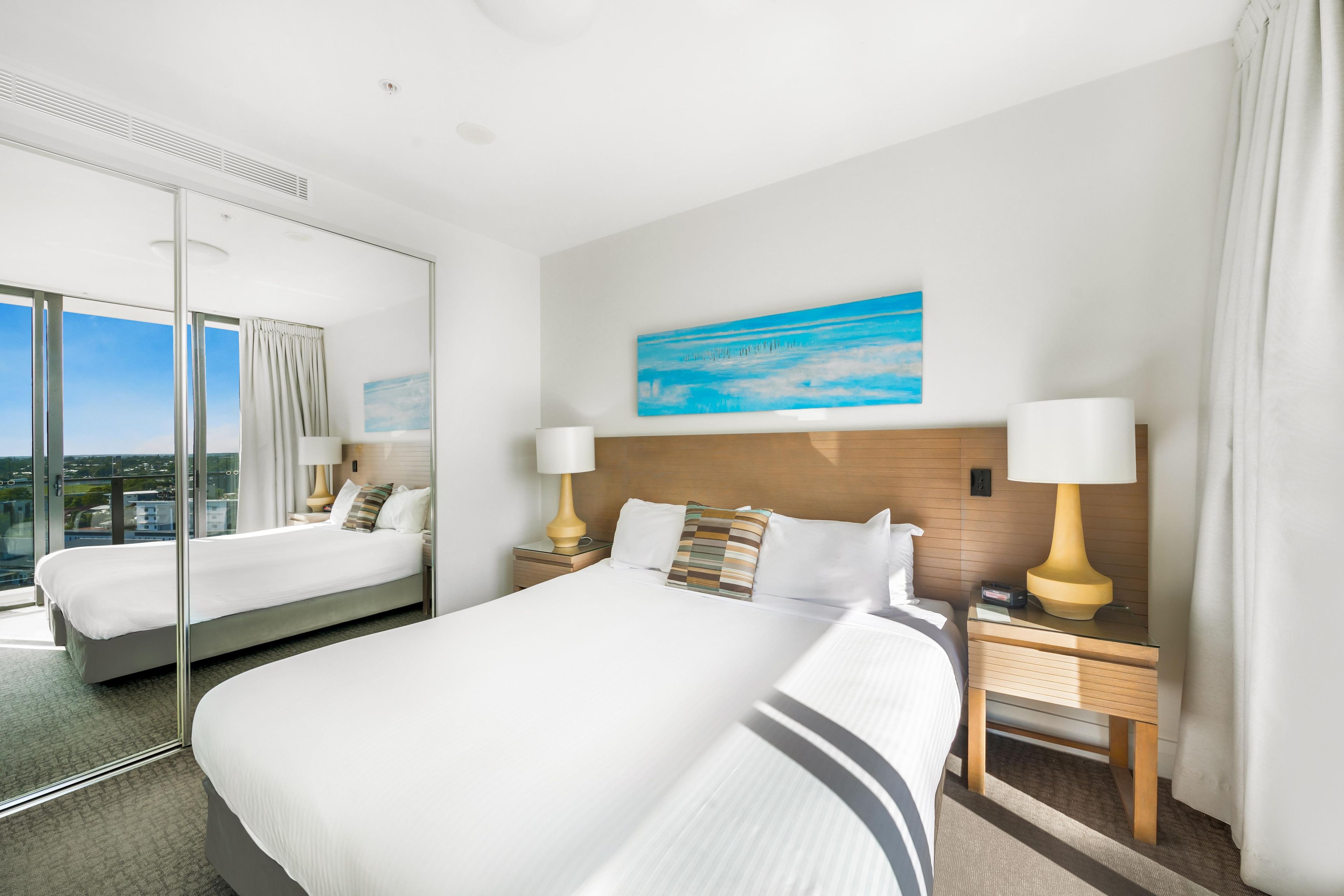 A hotel room with a large bed, two bedside tables with lamps, a painting on the wall, and a view through the window.