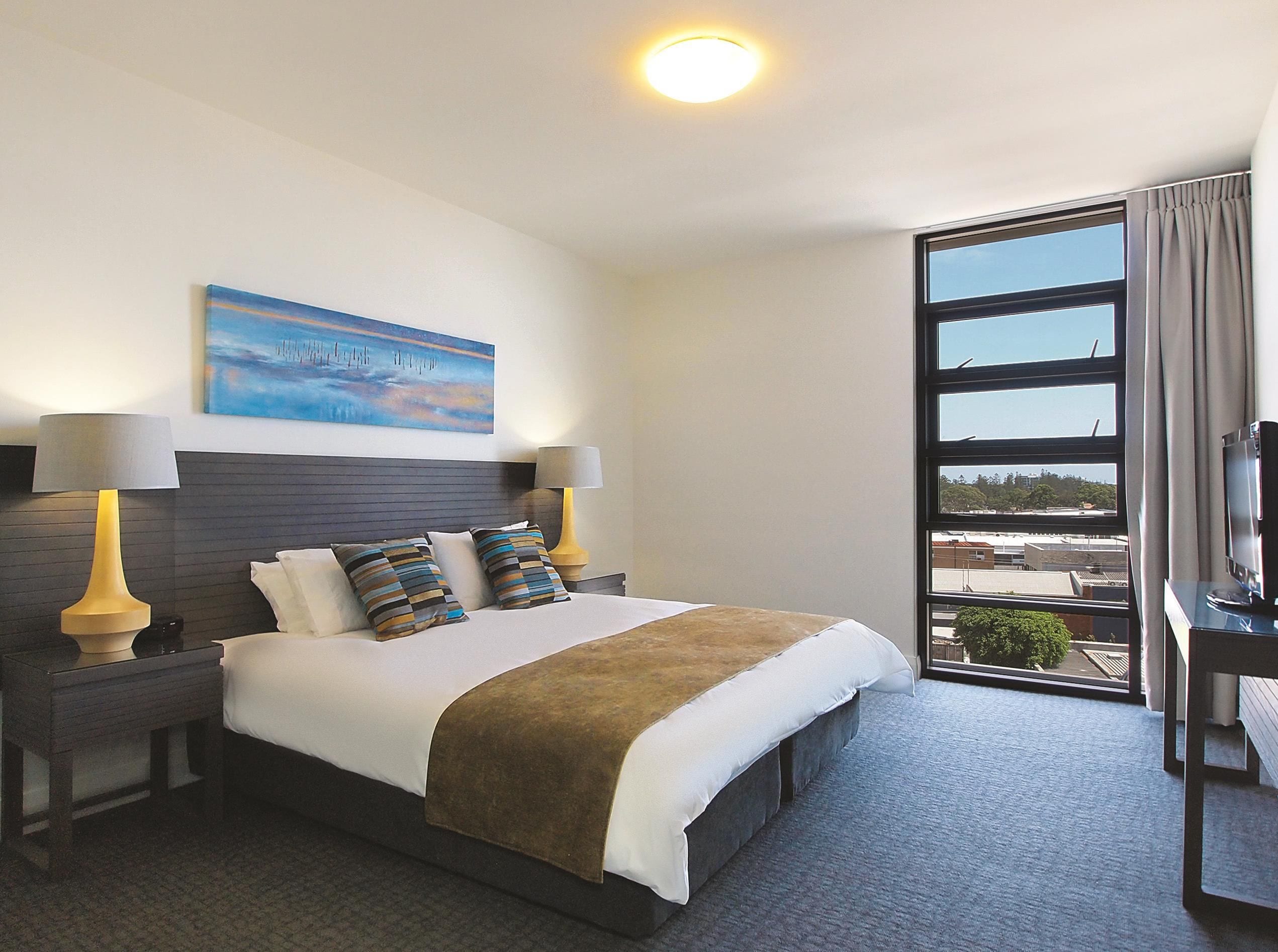 A hotel room with a bed, two bedside lamps, a painting on the wall, a television on a stand, and a large window with a view.