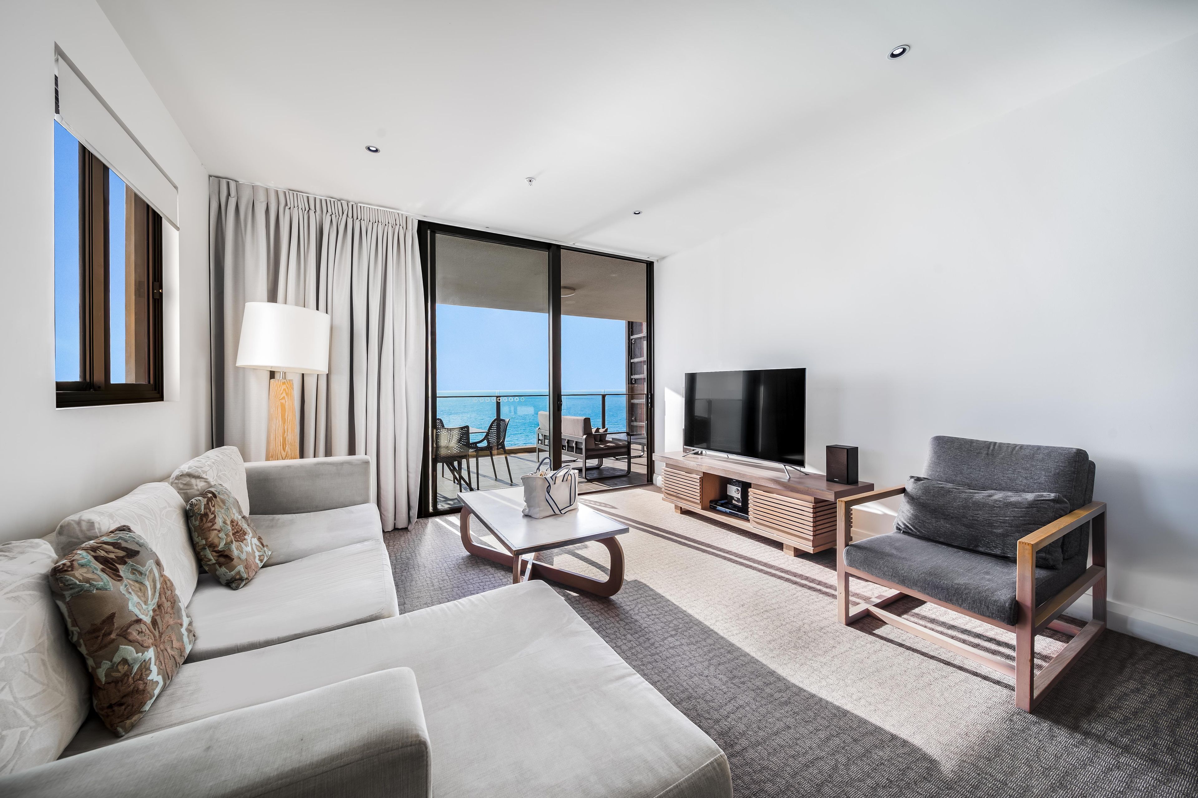 A modern hotel room with a sofa, a television, a coffee table, and a view of the sea.