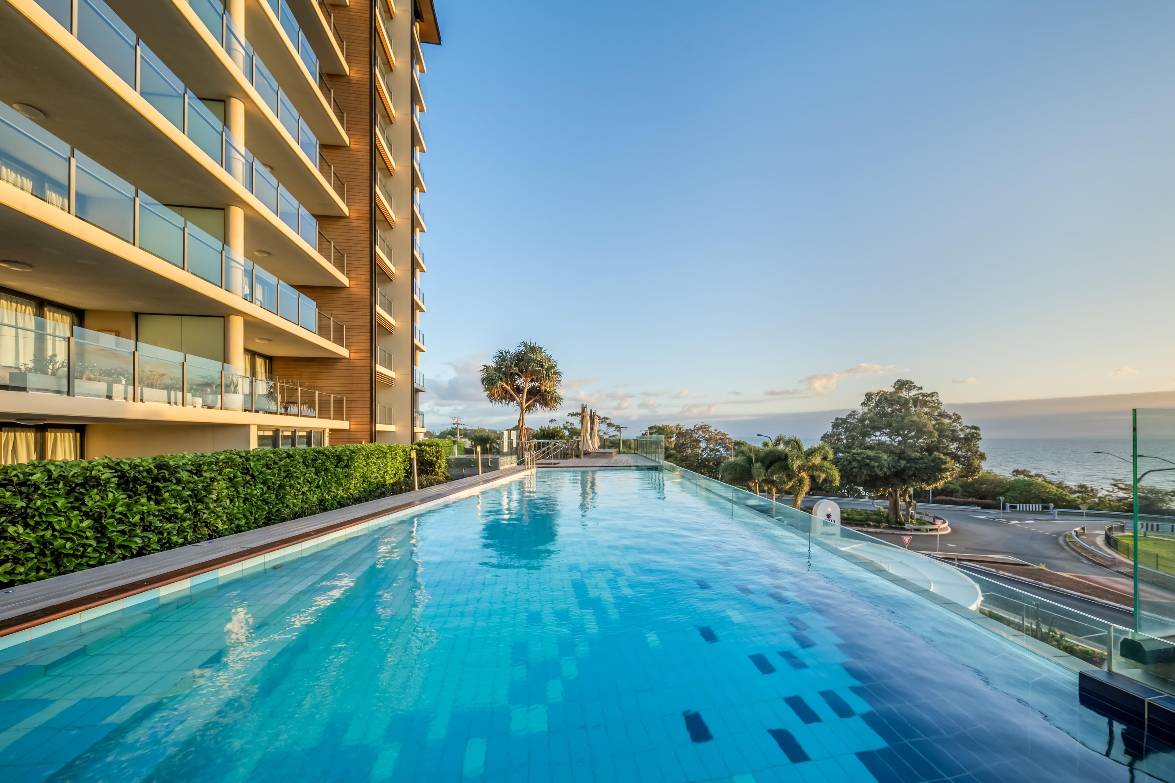 Una piscina junto a un hotel de varios pisos con balcones, rodeada de vegetación y vistas al mar.