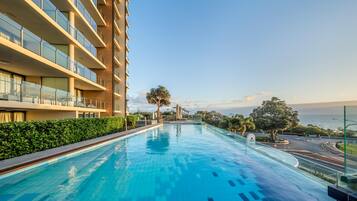 Una piscina junto a un hotel de varios pisos con balcones, rodeada de vegetación y vistas al mar.