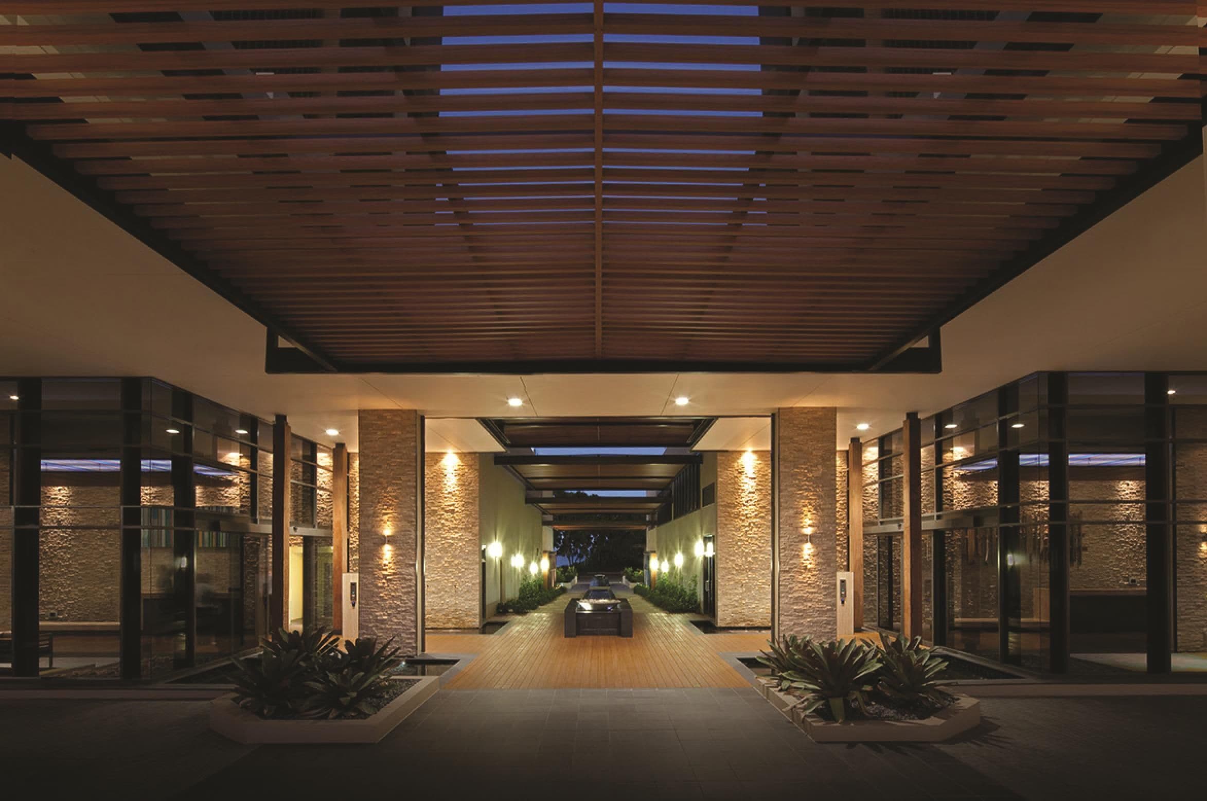 A modern hotel lobby with a wooden ceiling, stone walls, and a central pathway leading to the entrance.