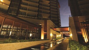 A modern hotel with multiple floors, outdoor seating areas, and a water feature in the foreground.