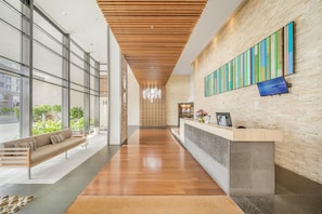 A modern reception area with a wooden ceiling, a reception desk, and a large window offering a view of greenery outside. - Oaks Redcliffe Mon Komo Suites (Redcliffe)