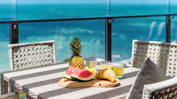 A wicker table with a wooden board holding a pineapple, watermelon slices, grapes, a croissant, and two glasses of juice, overlooking a clear blue ocean.