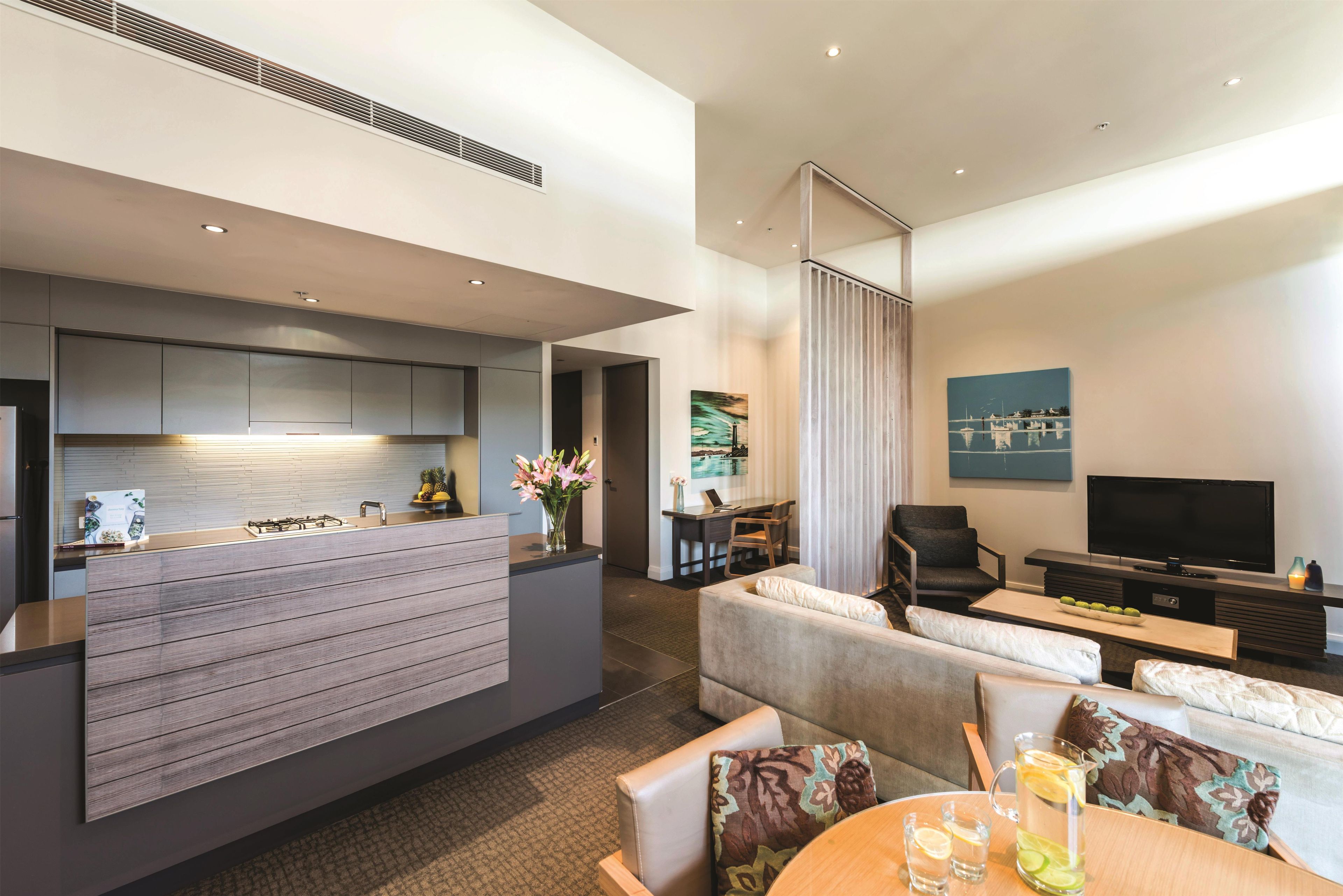 A modern hotel room with a kitchenette, a seating area with a sofa and armchairs, a flat-screen TV, and artwork on the walls.