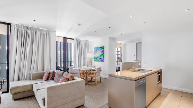 A modern living room with a sofa, a dining table, and a kitchen area with a sink and stove. - Oaks Redcliffe Mon Komo Suites (Redcliffe)