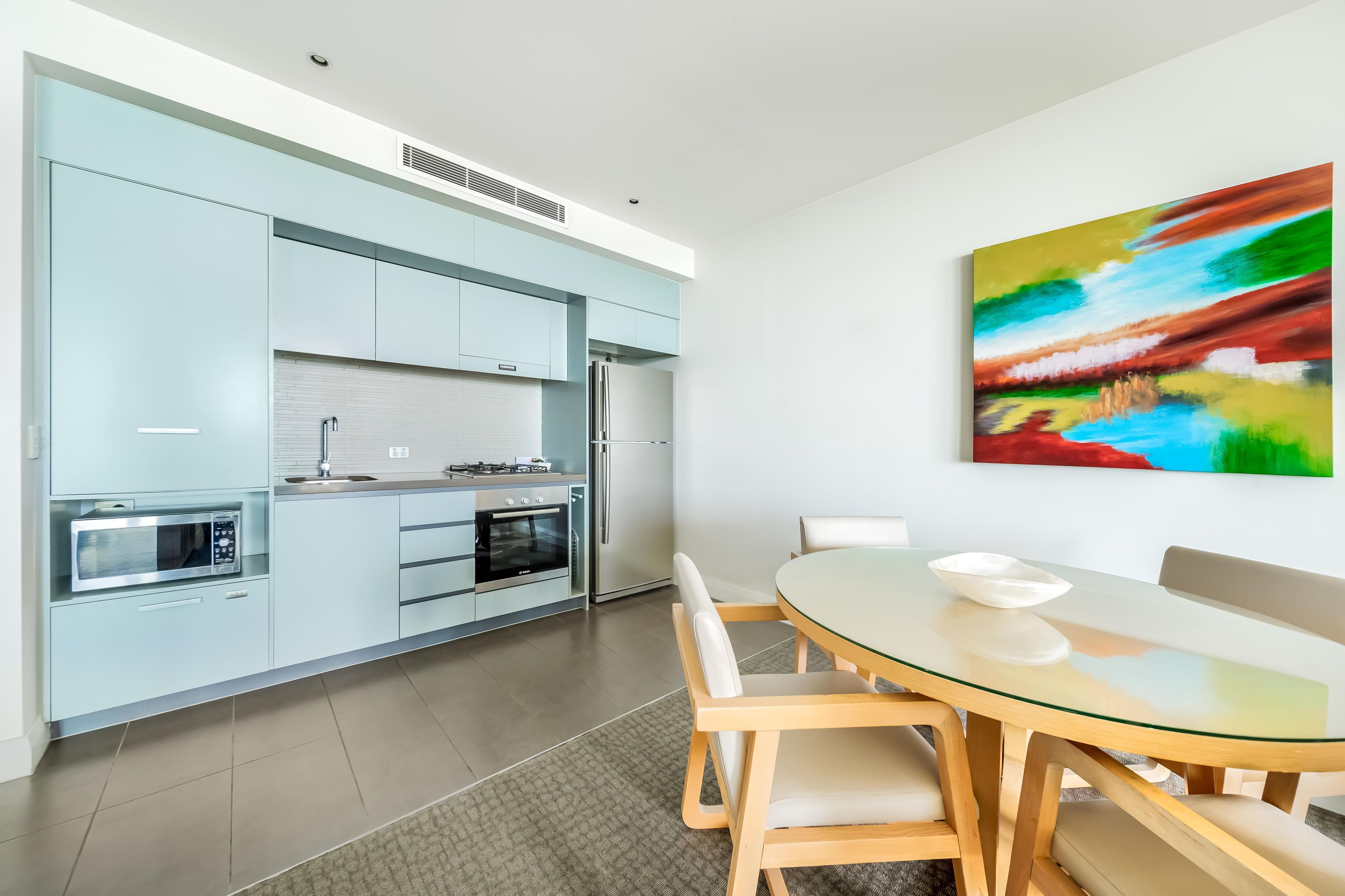 Una cocina moderna con electrodomésticos integrados, un comedor con mesa de vidrio y sillas de madera, y un cuadro abstracto y colorido en la pared.