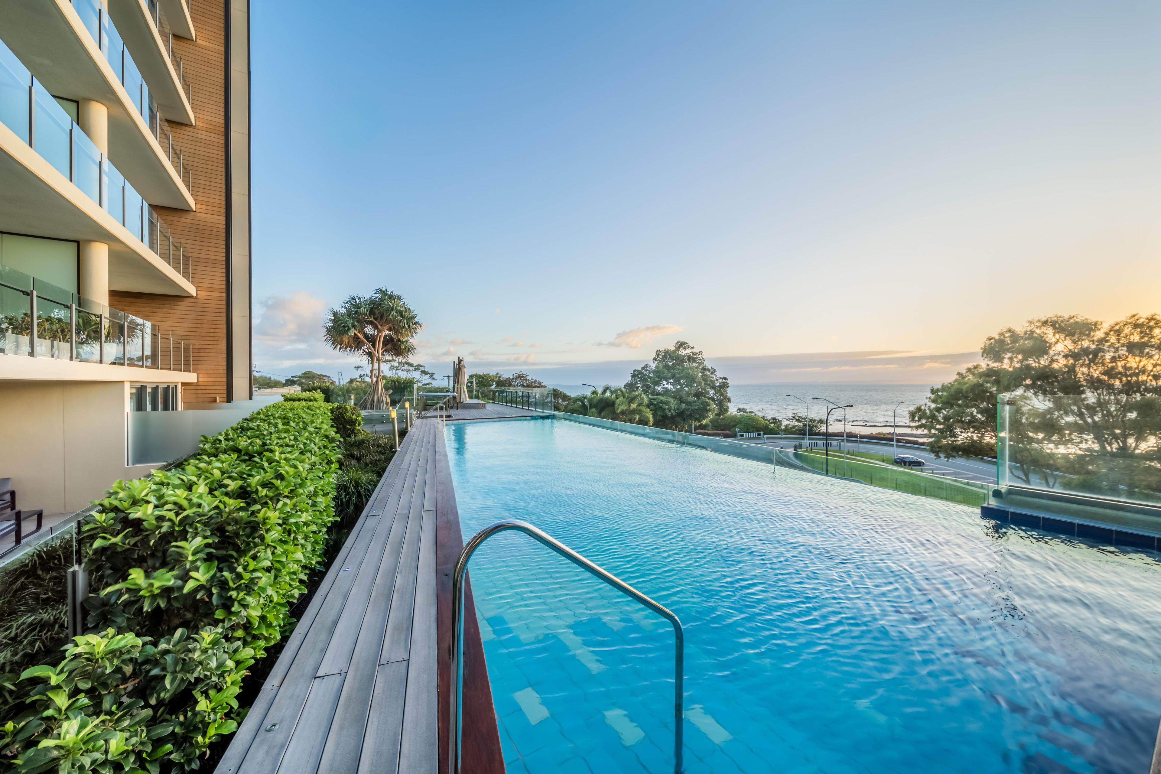 A swimming pool with a wooden deck, surrounded by greenery, and overlooking a body of water.