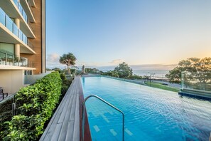 A swimming pool with a wooden deck, surrounded by greenery, and overlooking a body of water. - Oaks Redcliffe Mon Komo Suites (Redcliffe)