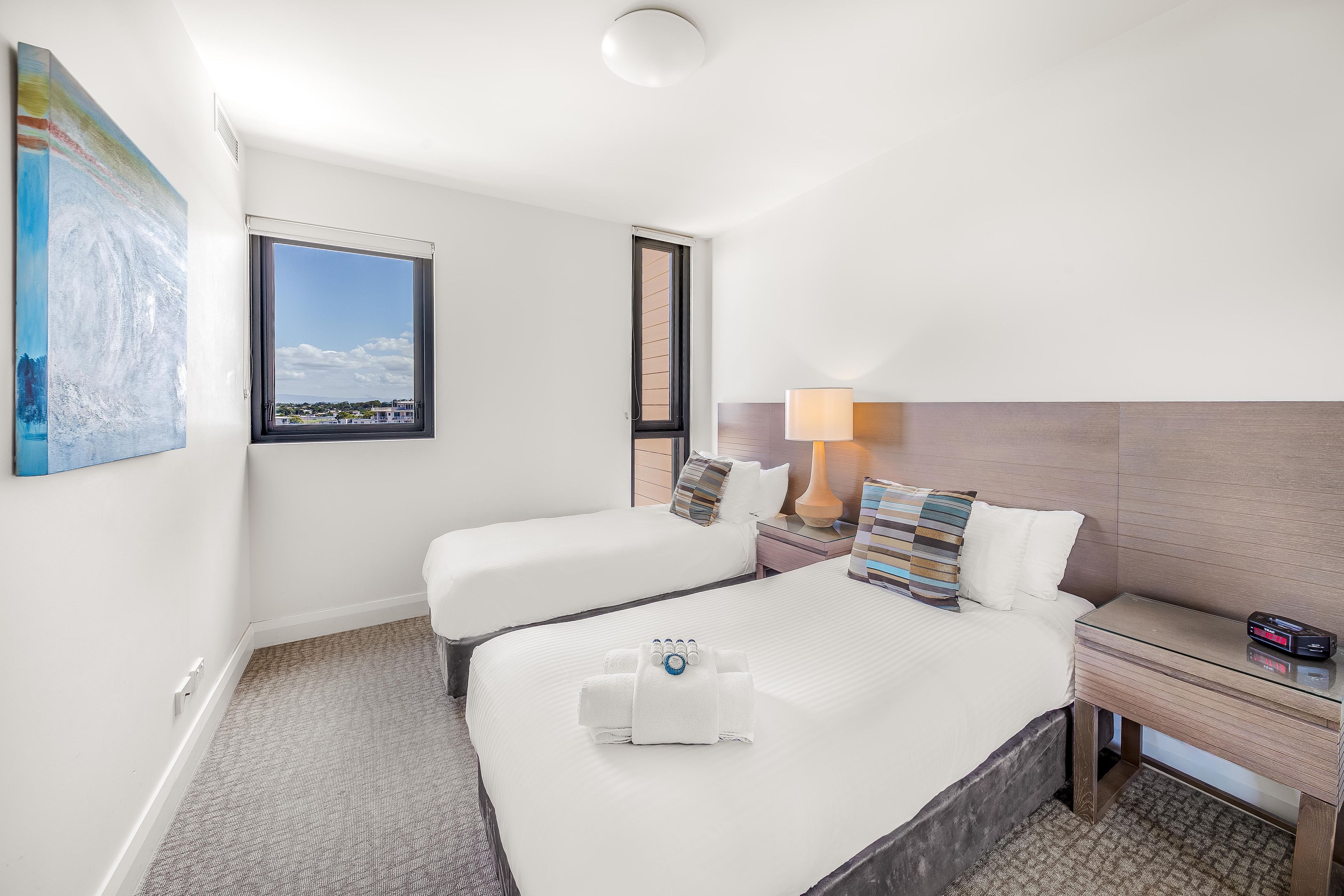A hotel room with a bed, two bedside tables, a lamp, a clock, and a painting on the wall.