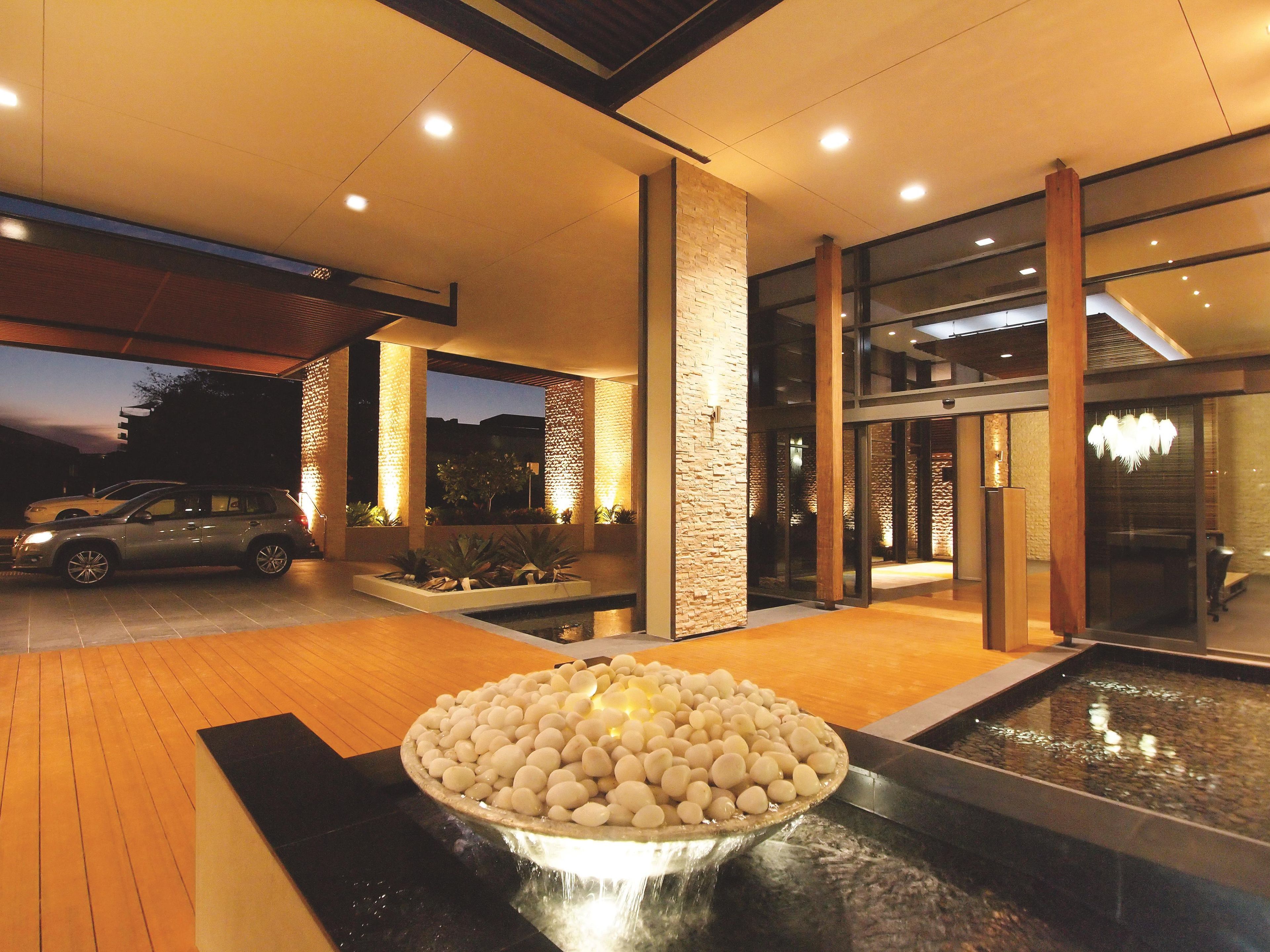A modern building exterior with a car parked outside, a water feature with stones, and a well-lit entrance.