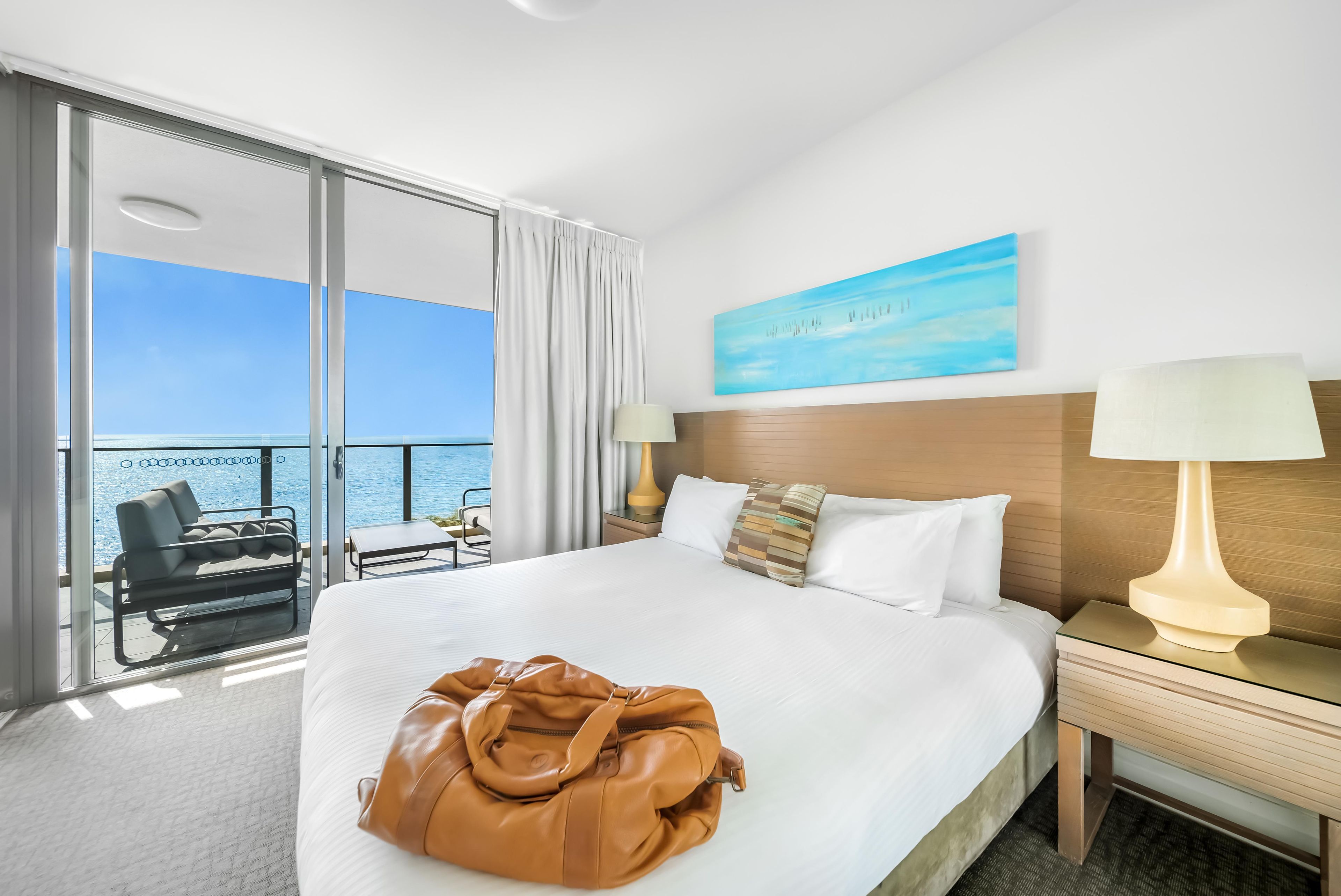 A hotel room with a bed, a nightstand, a lamp, a view of the sea, and a painting on the wall.