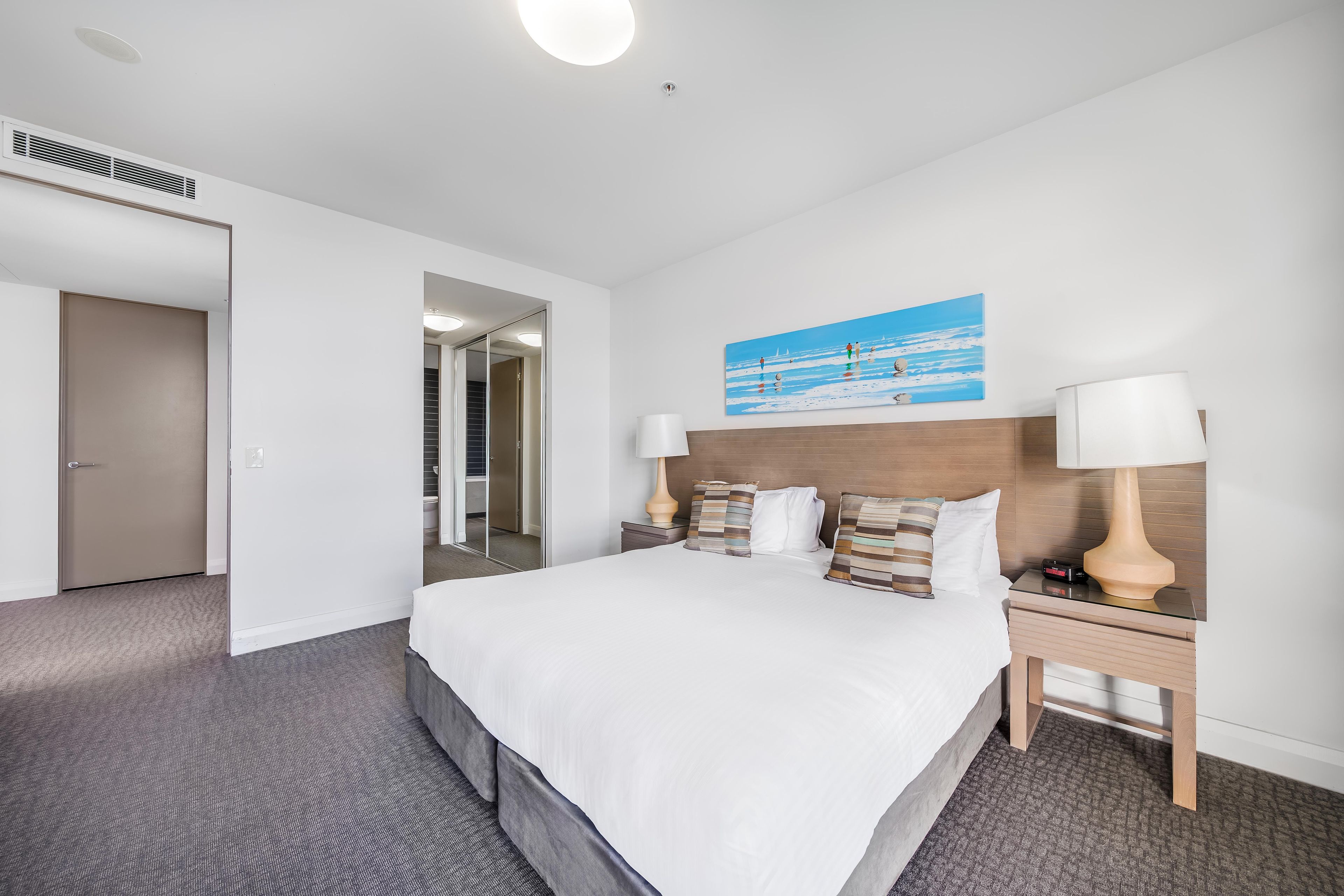 Habitación de hotel con una cama grande, dos mesitas de noche, una mesita auxiliar, un cuadro y una puerta que da a un baño.