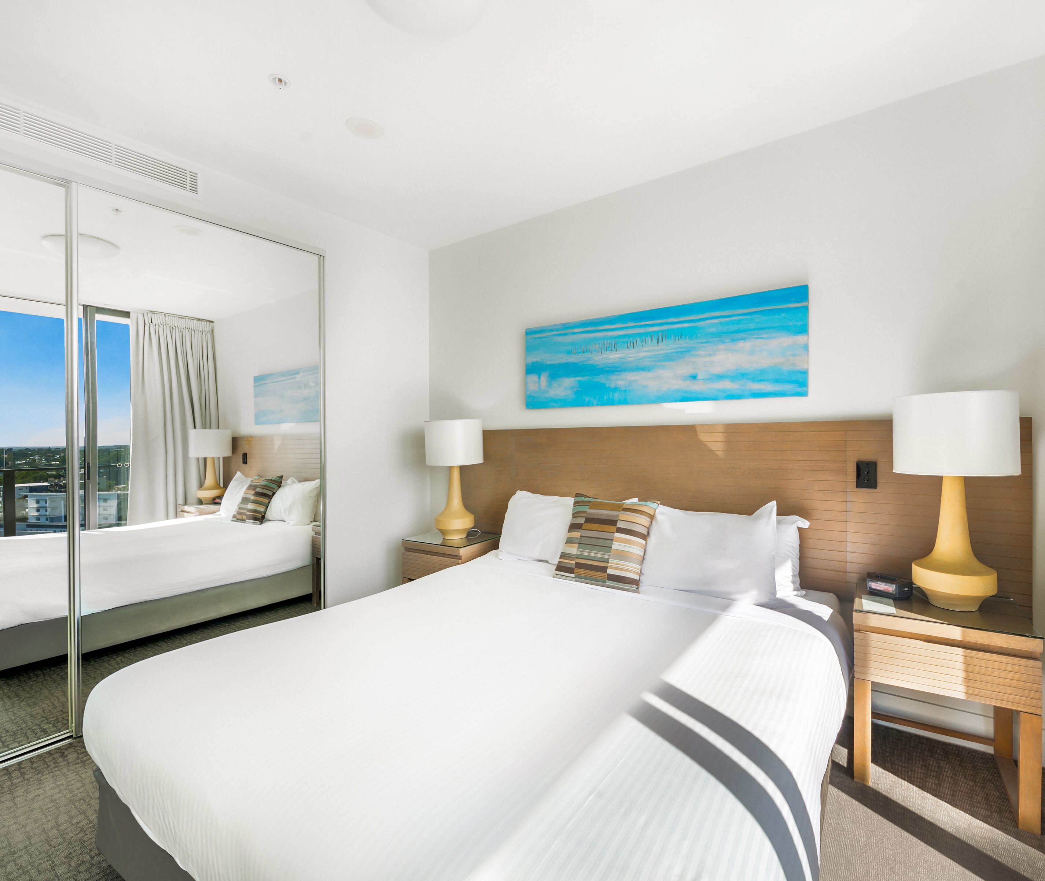 Quarto de hotel com uma cama grande, duas mesas de cabeceira com luminárias, um quadro na parede e uma janela ampla com cortinas.