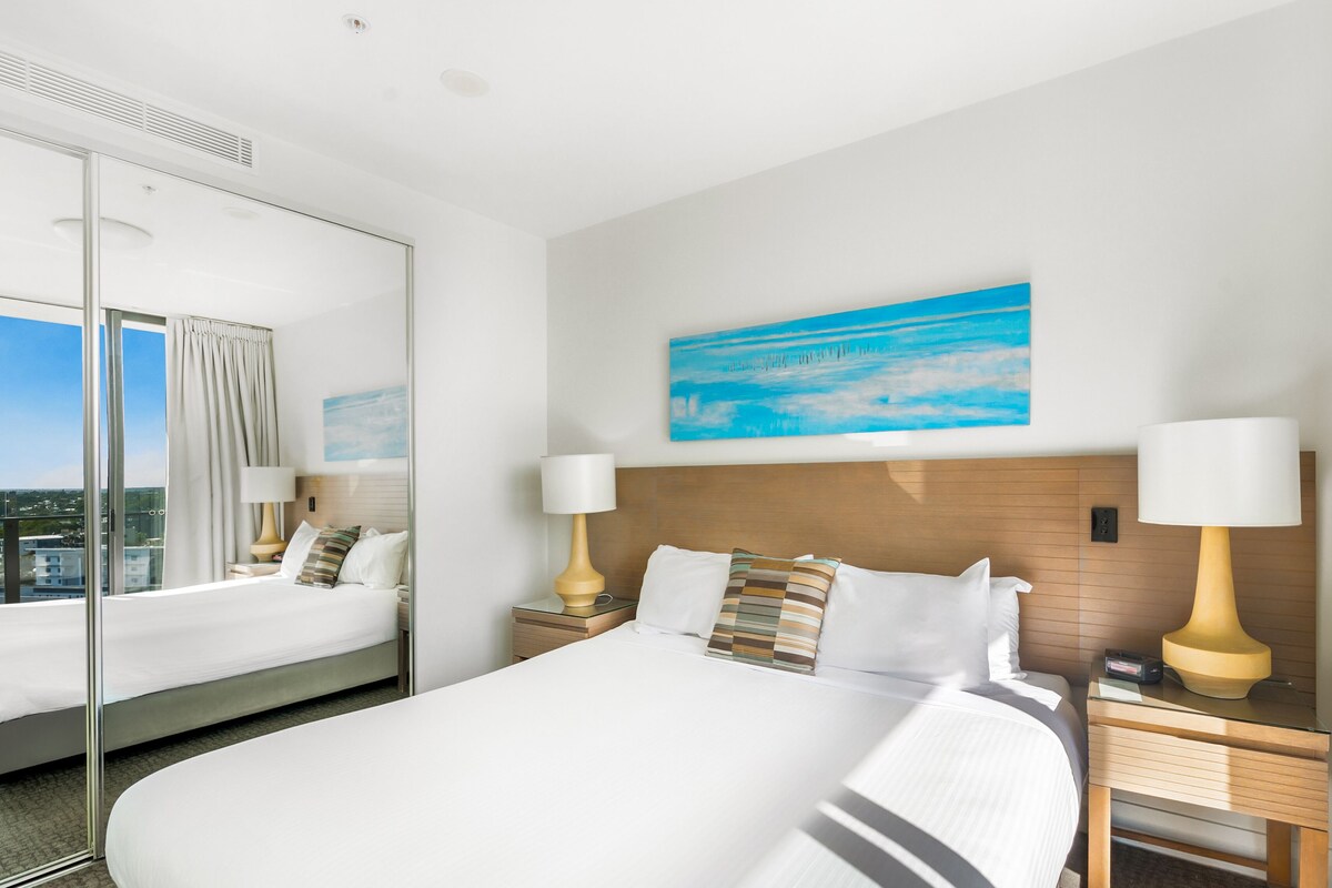 A hotel room with a large bed, two bedside tables with lamps, a painting on the wall, and a large window with curtains.
