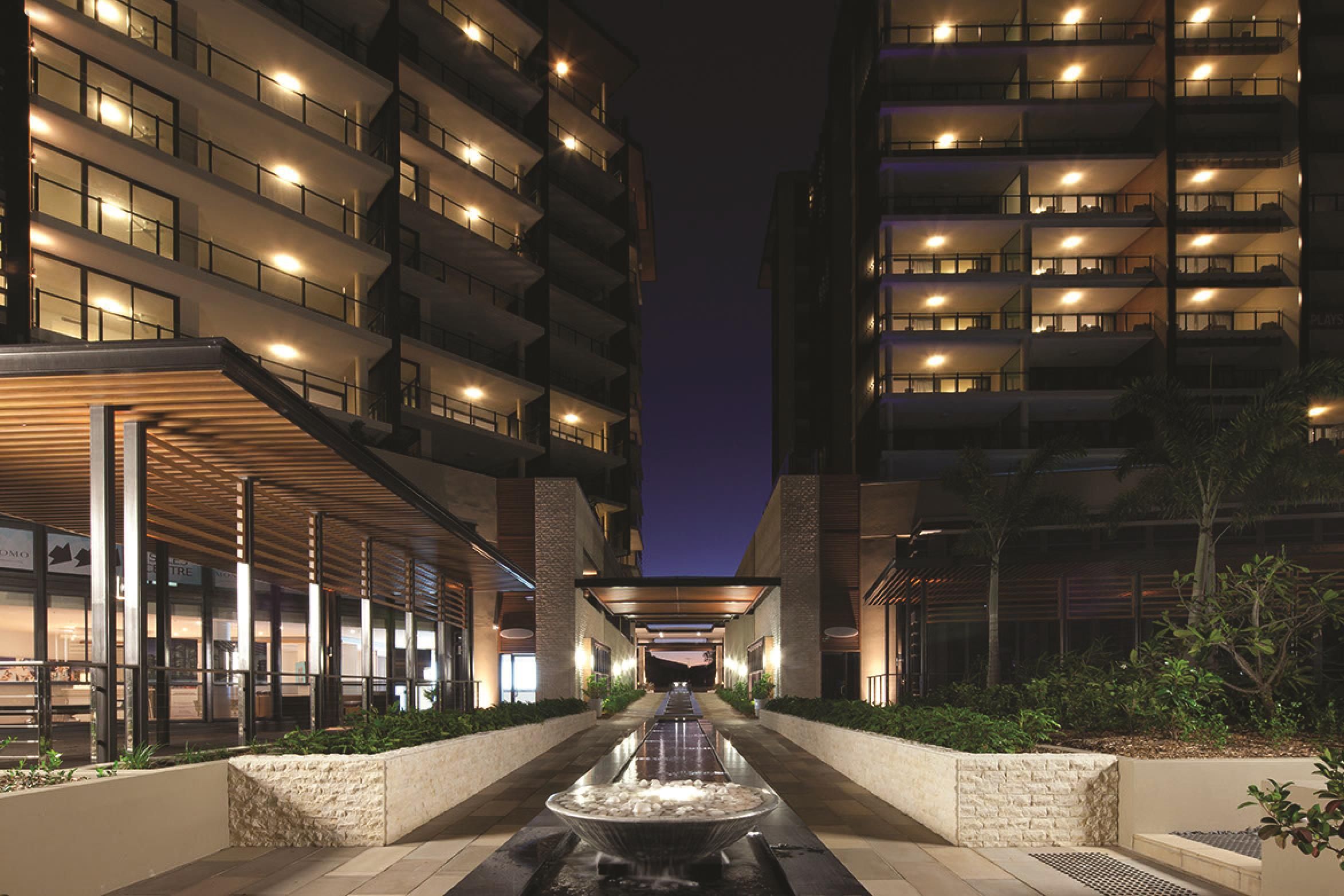 A nighttime view of a hotel entrance with a central water feature, multiple floors with illuminated windows, and a covered walkway on either side.